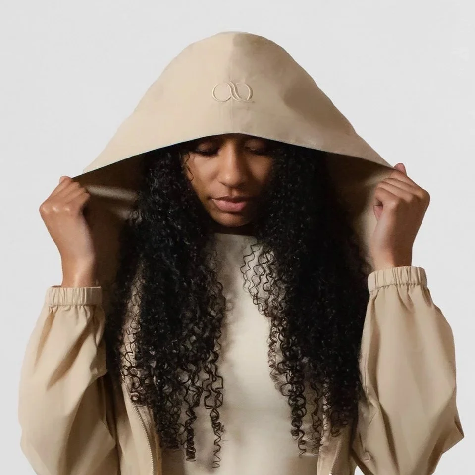 A woman with long curly hair wearing a beige hooded jacket with her hands holding the hood, looking downward with her eyes closed.