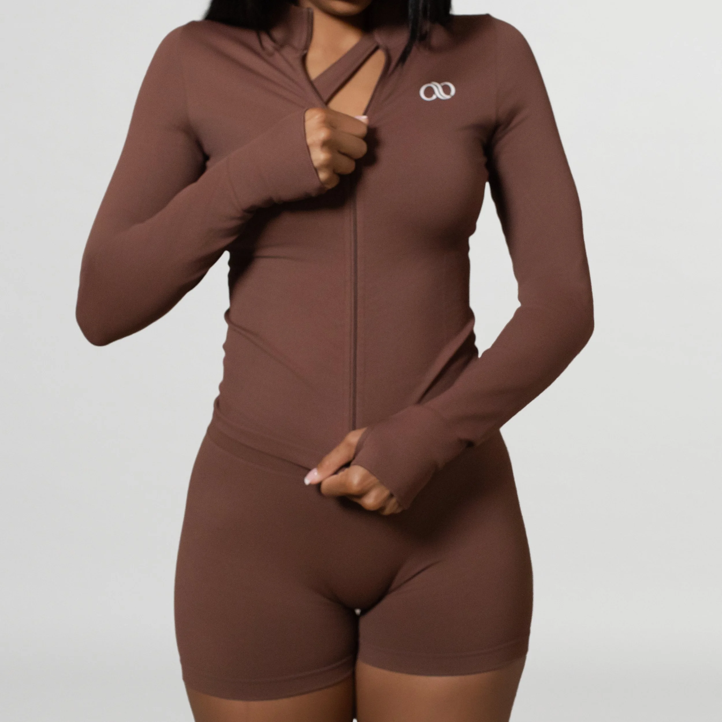 A woman is wearing a matching brown athletic outfit consisting of a zip-up jacket and high-waisted shorts, standing against a plain white background.