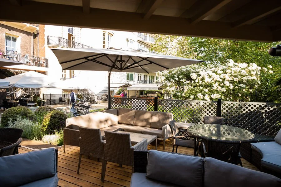 An outdoor patio with wicker and cushioned seating, large umbrellas, and a garden with flowering bushes, in front of apartment buildings.