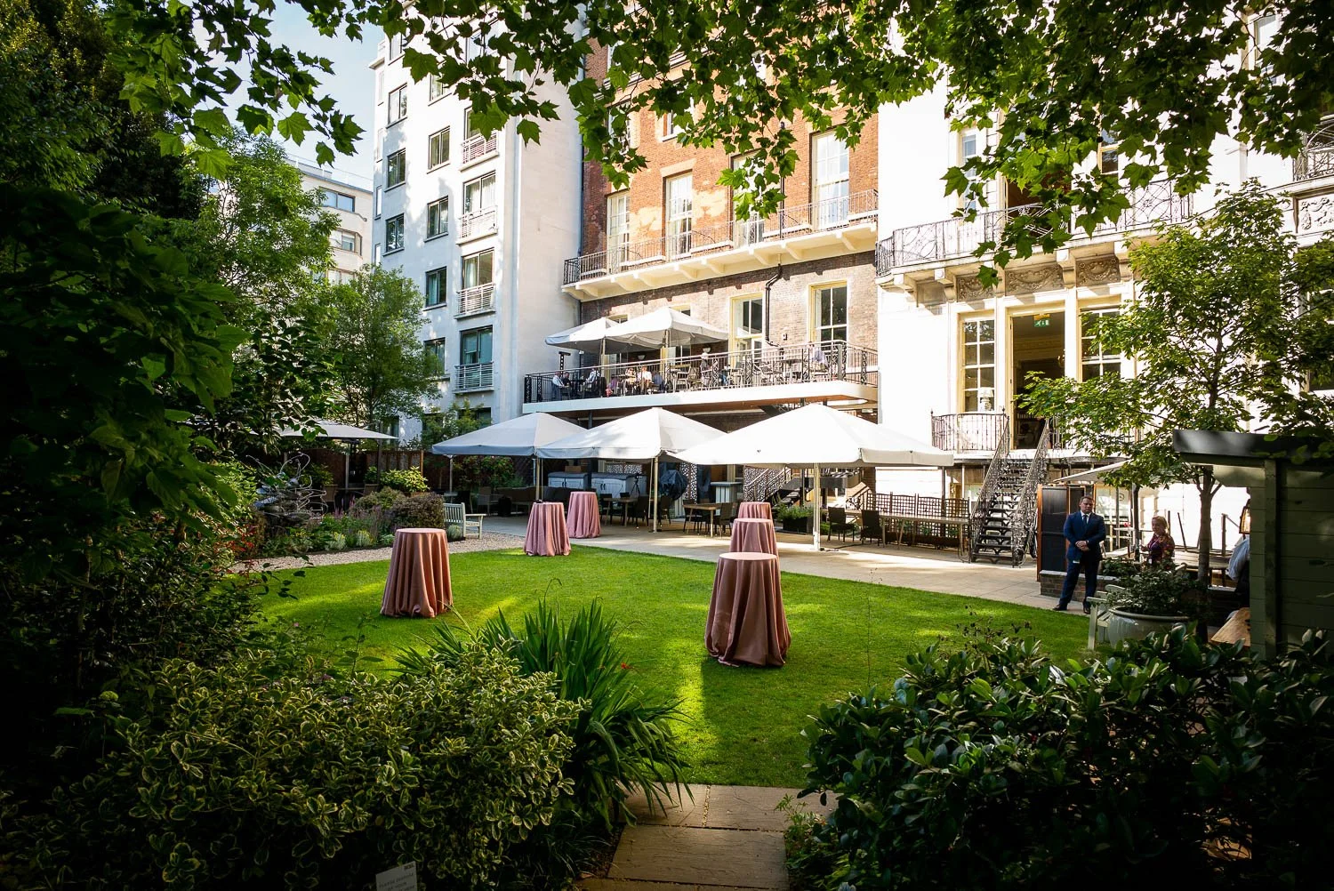 An outdoor garden patio area with tall tables draped in pink cloths, surrounded by lush greenery, white umbrellas shading seating areas, and a multi-story building with balconies and large windows in the background. Two people are standing and conversing near the building, and there are other seated guests and staff visible.