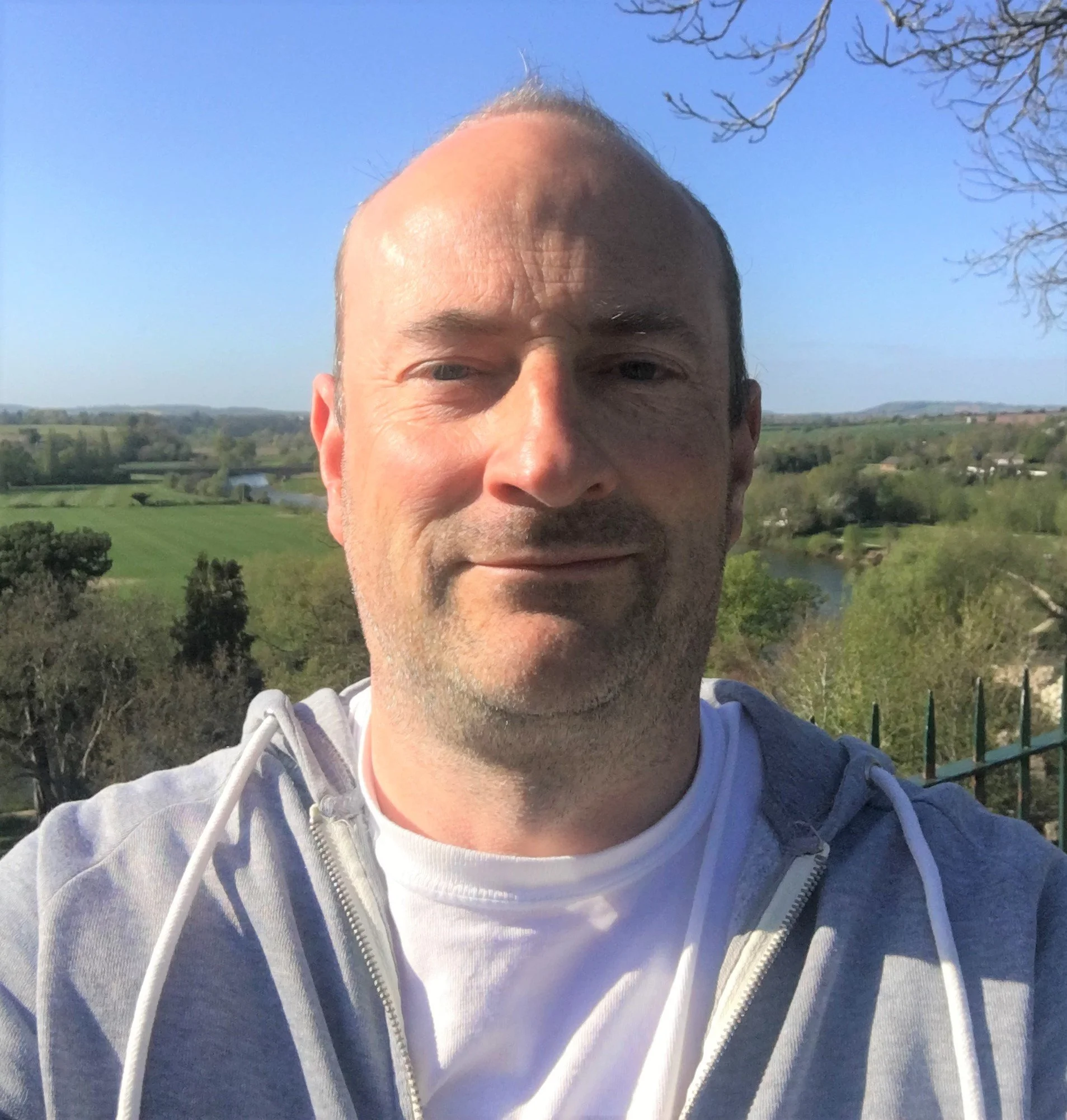 Person taking a selfie outdoors with a scenic background of green fields and a blue sky.