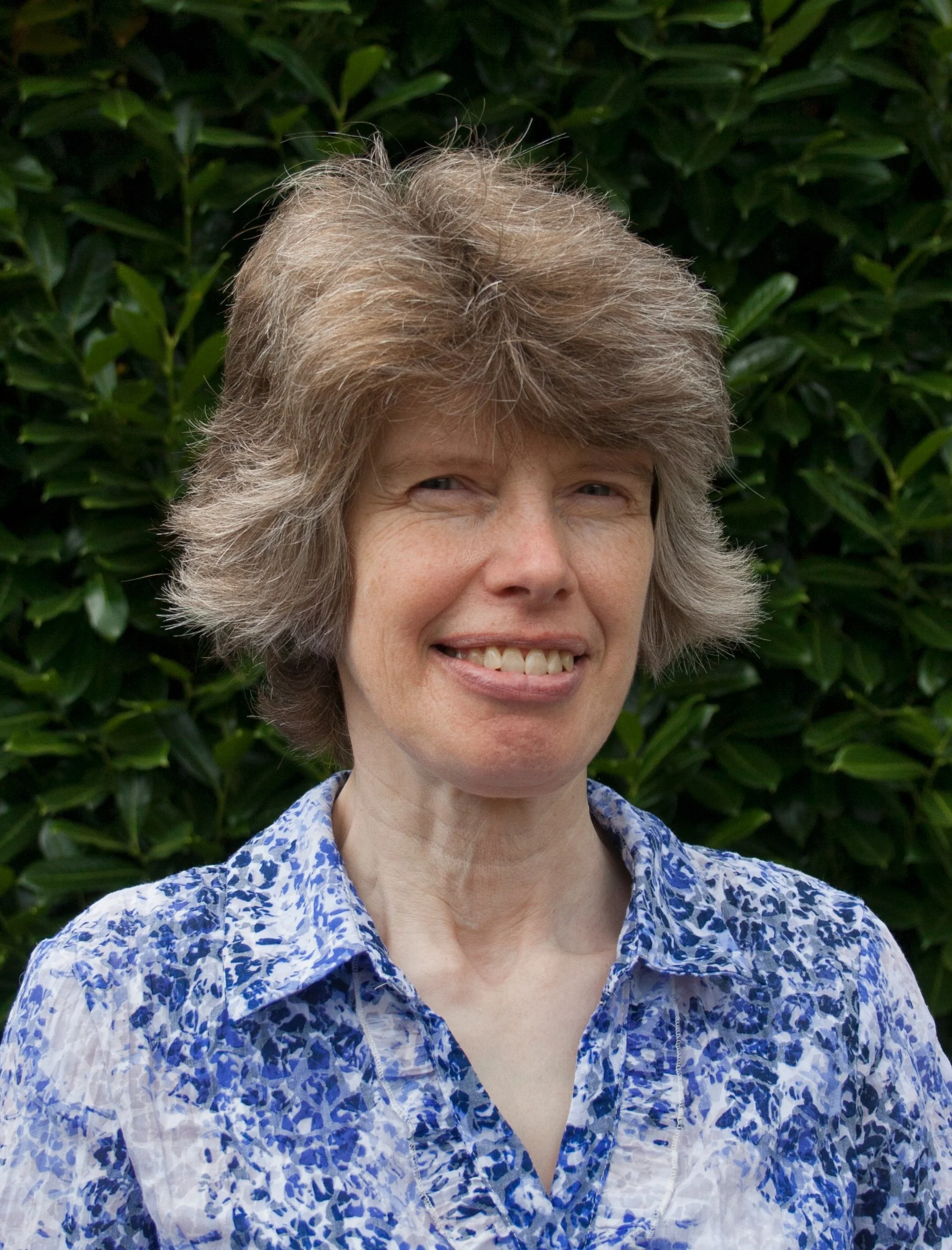 Person smiling with short, gray hair, wearing a floral shirt, standing in front of green foliage.