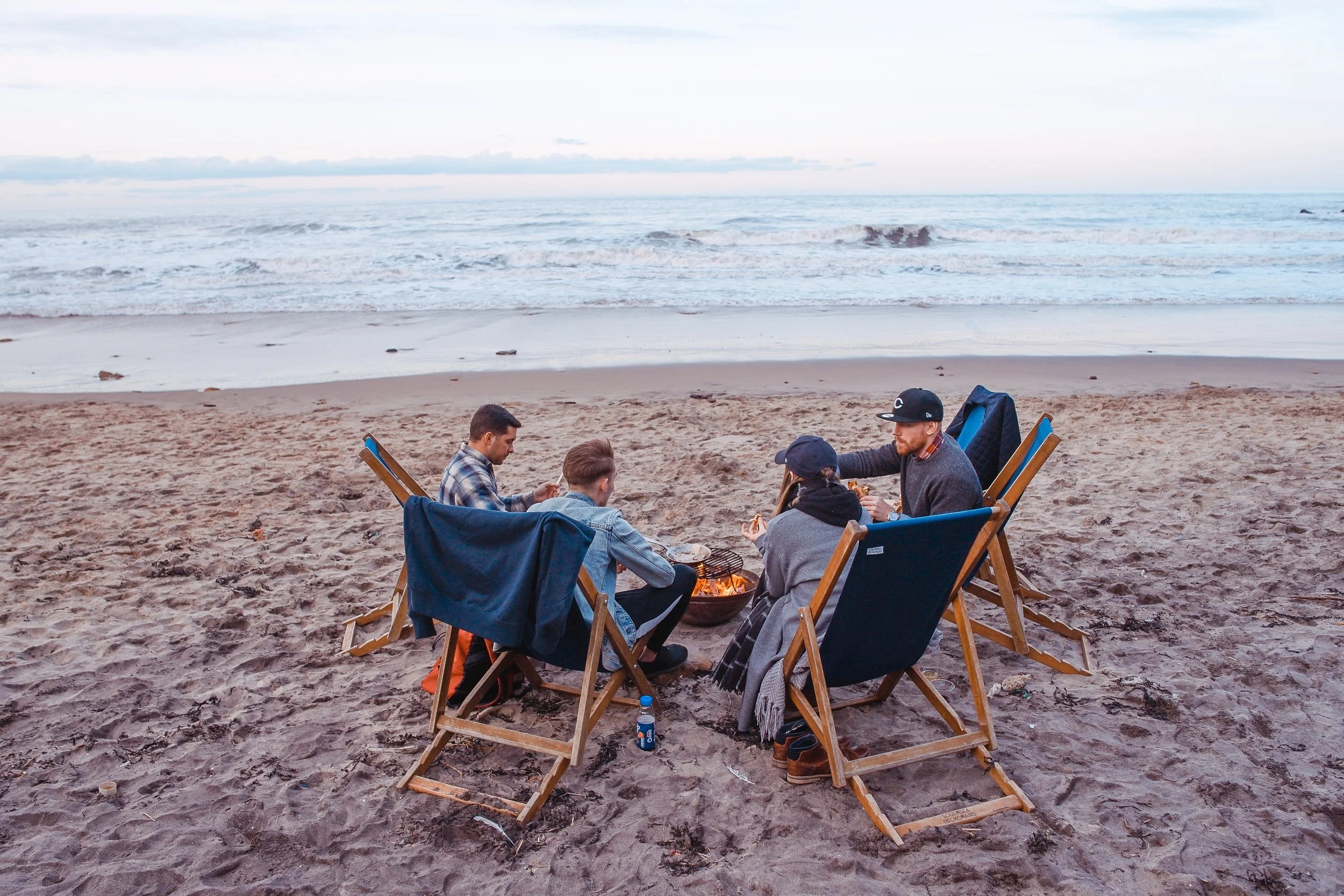 Group of people sitting around a beach campfire in foldable chairs, with ocean waves in the background.