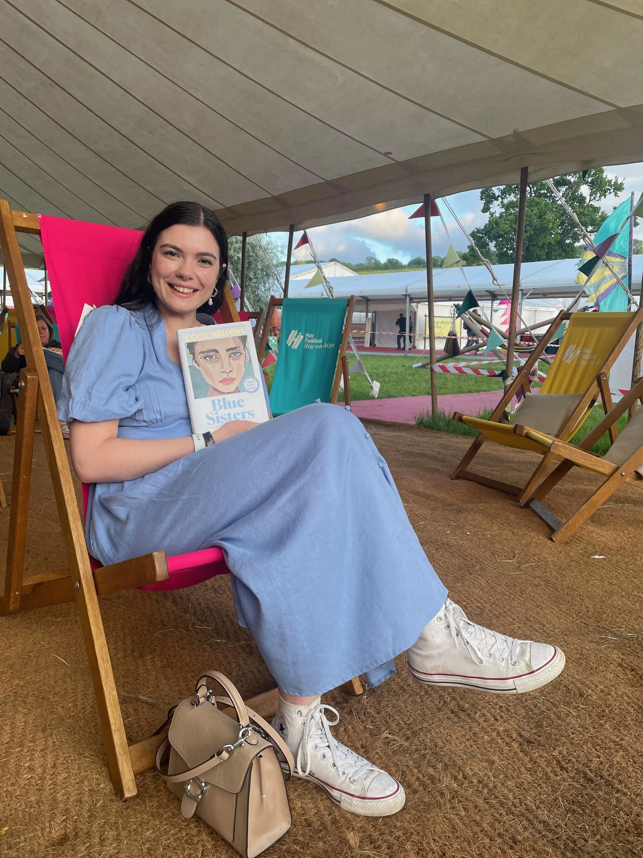 Lulu wears a blue dress, sitting on a deck chair under a festival tent, smiling at the camera and holding a copy of Blue Sisters by Coco Mellors