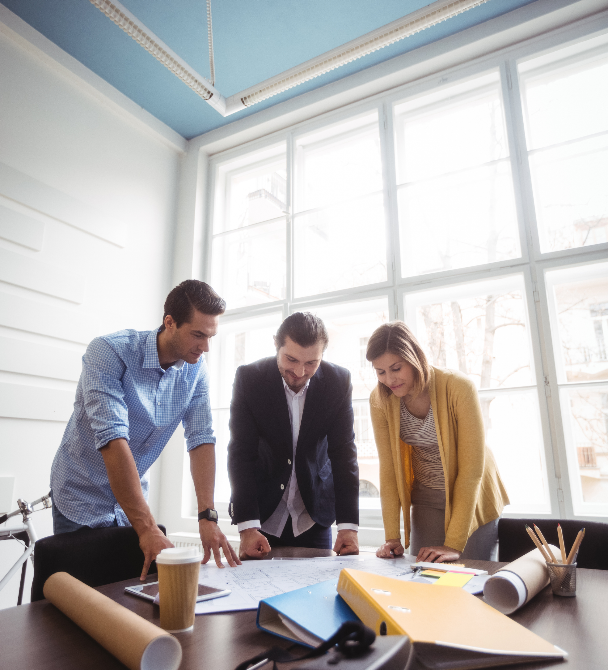 Architect firm meeting happening indoors in front of a large window three entrepreneurs looking at blueprints