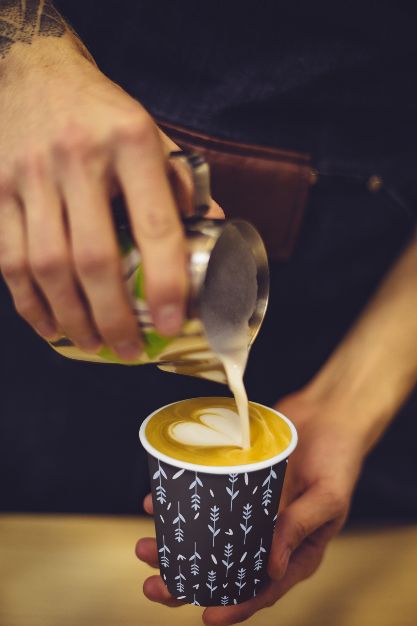 Person pouring milk into a cup of coffee with latte art in a black and white patterned paper cup.
