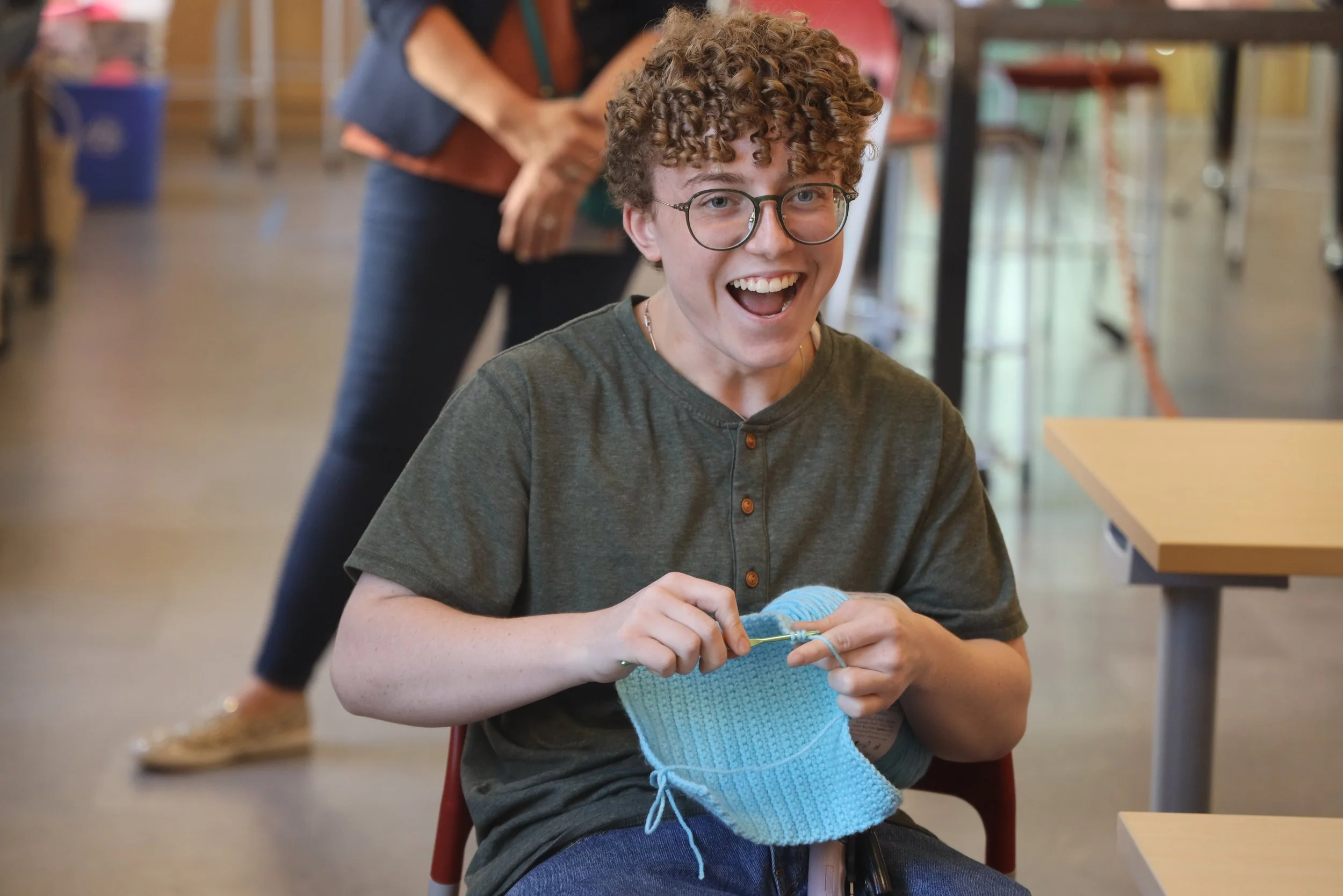 A young person with curly hair and glasses smiling while knitting a blue piece of fabric in a bright indoor space.