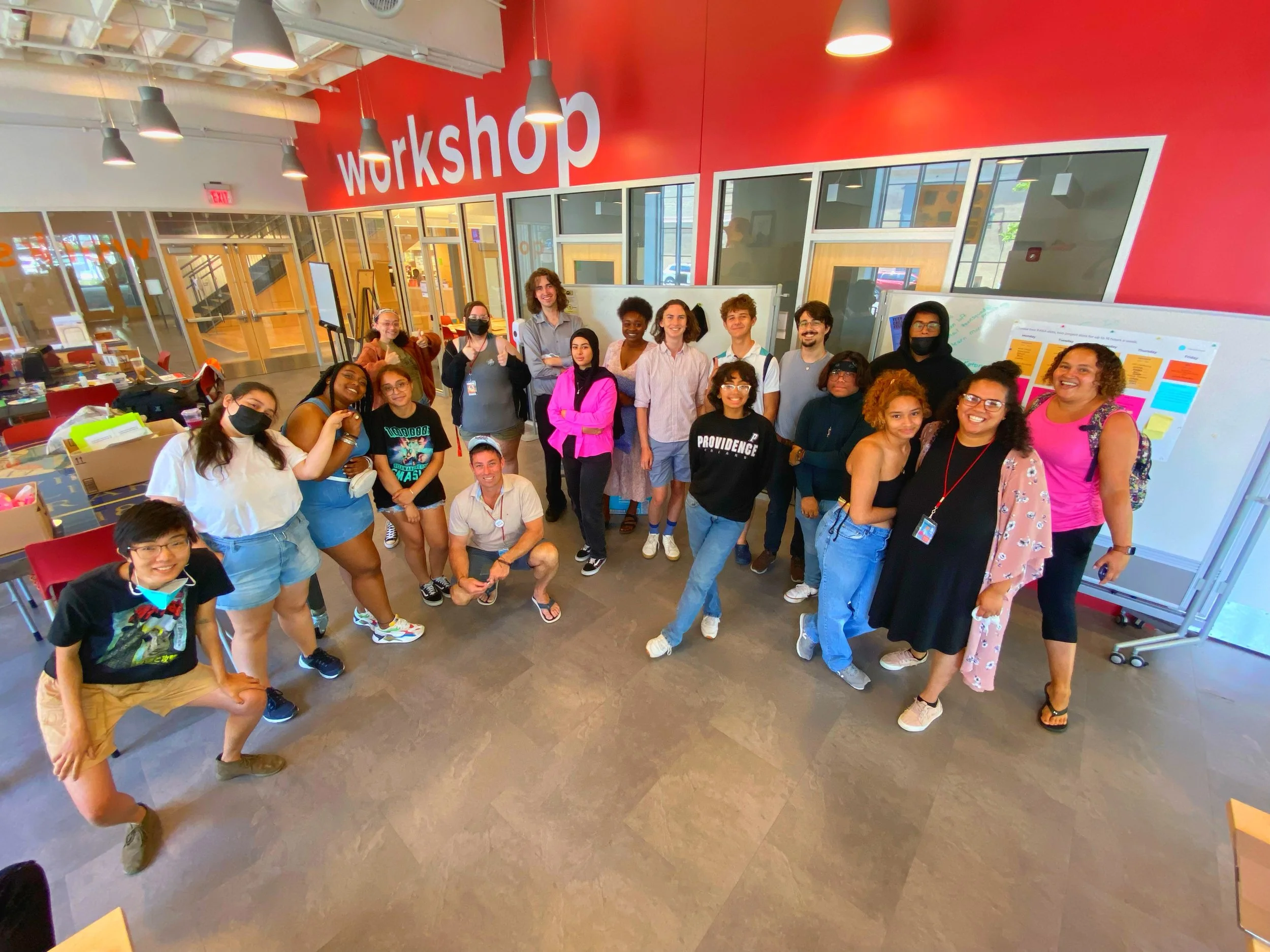 Group of diverse people gathered in a workshop space, some smiling and making gestures, with a red wall that says 'workshop' behind them