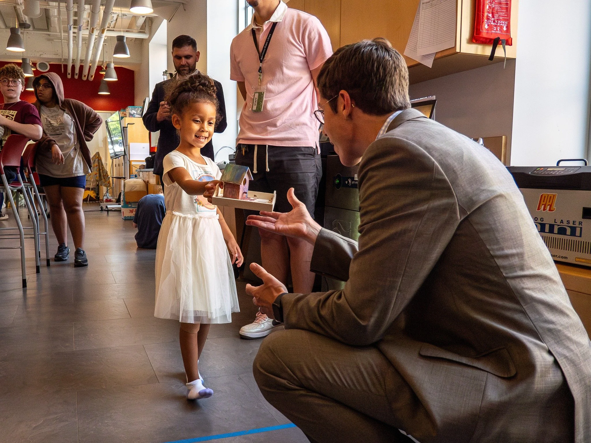 A young girl in a white dress receiving a small gift from a man in a gray suit who is crouching down and talking to her at an indoor event with several people in the background.
