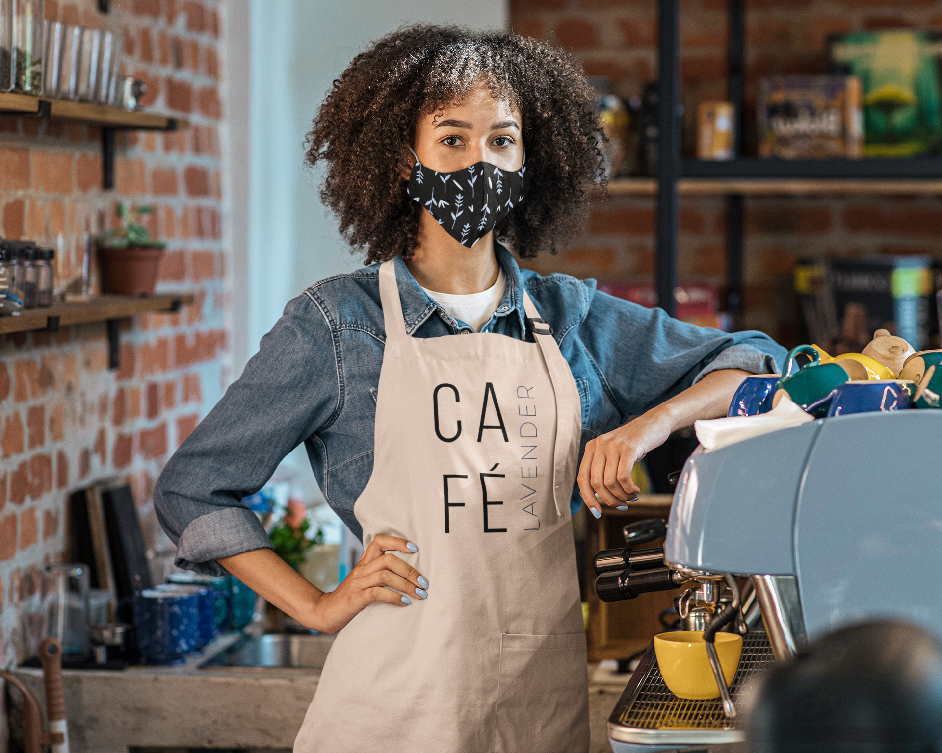 A woman wearing a denim shirt, apron, and face mask stands behind a coffee machine in a cafe, with shelves of supplies and exposed brick wall in the background.