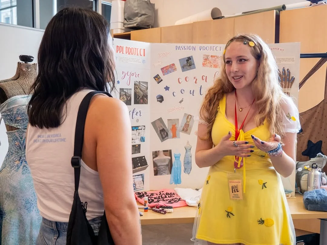 Two women are having a conversation at an event or workshop in front of a presentation board titled 'PASSION PROJECT'. One woman has long dark hair, is wearing a sleeveless white top and has a black bag; the other woman has long blonde hair, is wearing a yellow dress with small embroidered insects, and has a red lanyard with an ID badge.