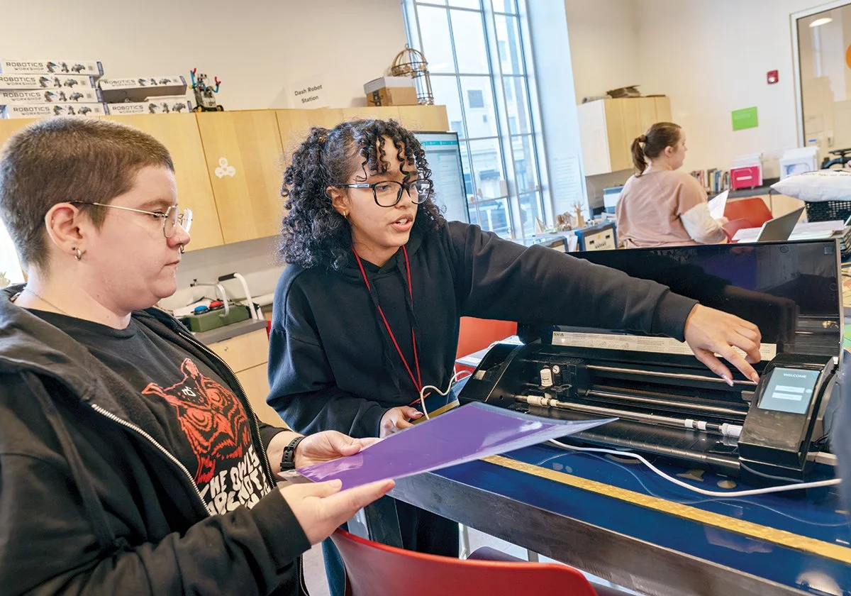Two women working on a laser cutter in a classroom or workshop. One woman is holding a purple sheet and the other is operating the machine while looking at it.