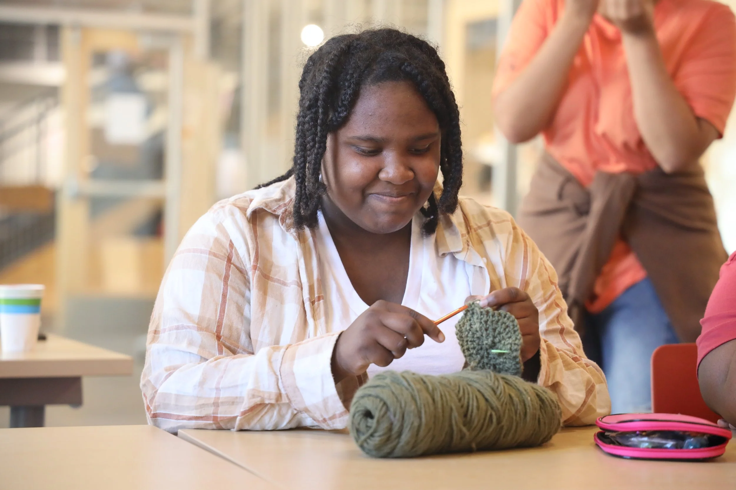 A young woman with dark, curly hair knitting with green yarn in a cozy indoor space, with other people and a shelving area in the background.