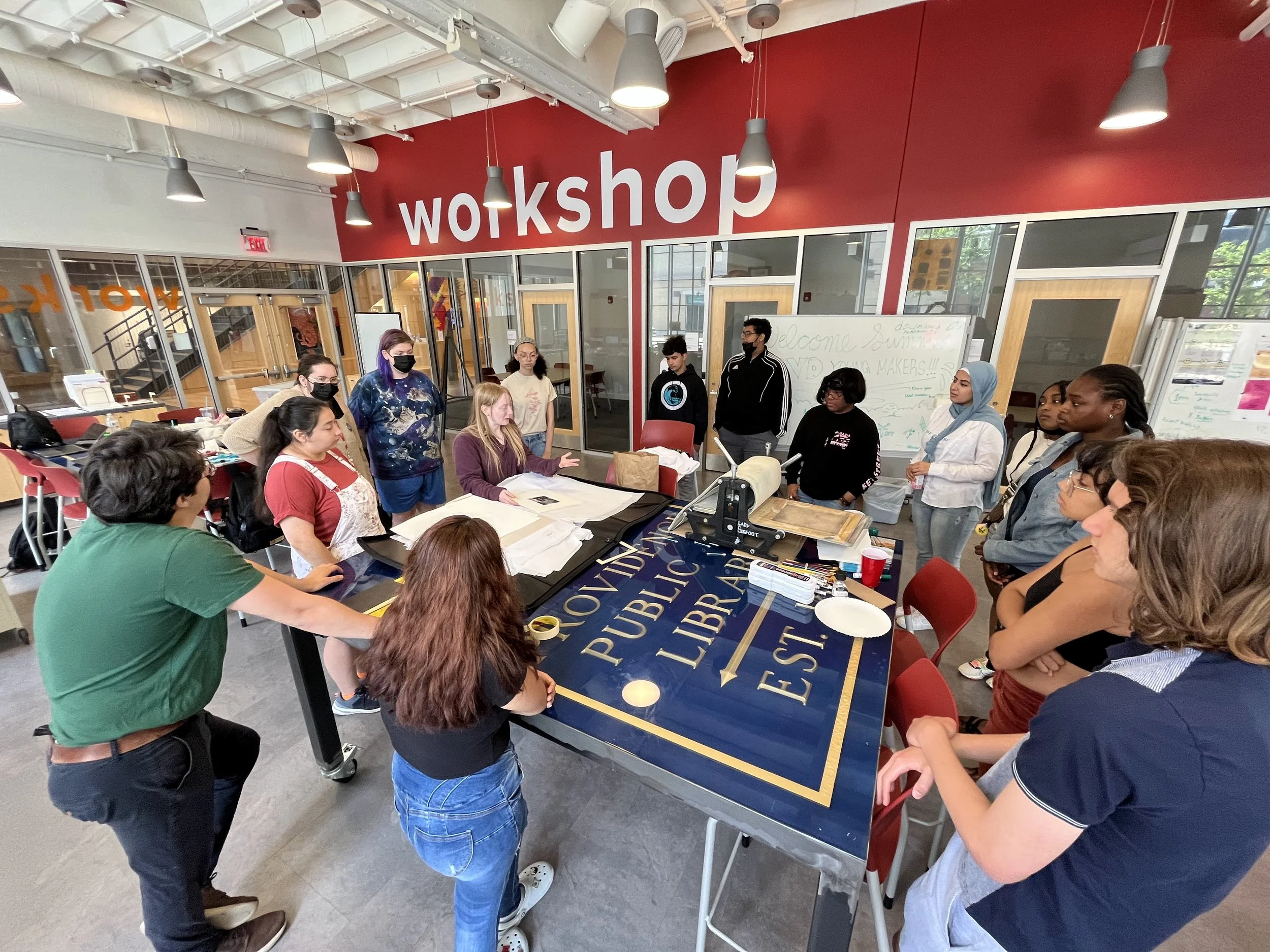 A group of people gathered around a large conference table in a workshop room, listening to a woman in a purple sweatshirt who is speaking. The room has red walls, a whiteboard with writing, and a sign that says 'wokshop' on the wall.