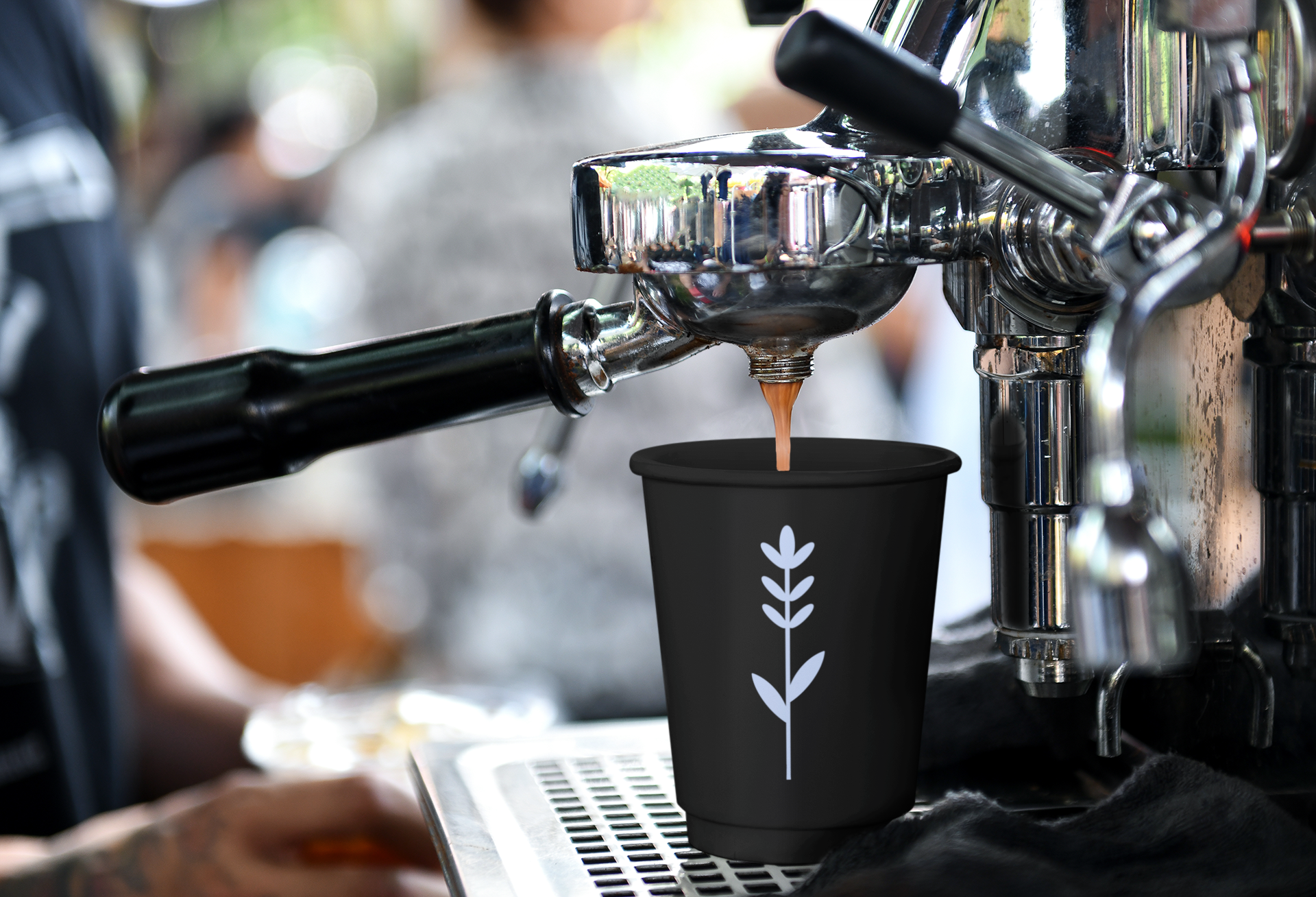 Close-up of a espresso machine brewing coffee into a black cup with a white leaf design, in a cafe setting with blurred people in the background.