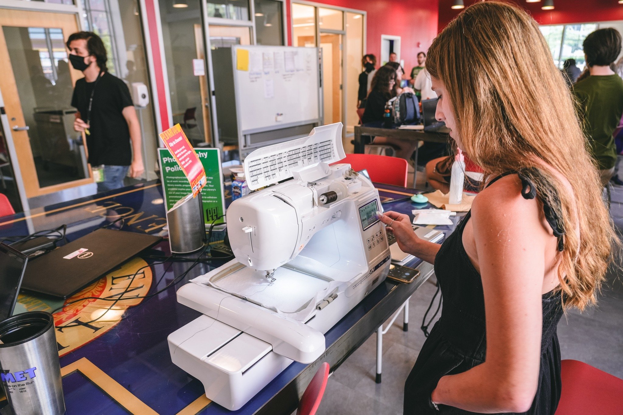 A woman operating a sewing machine at a table in a busy indoor setting, with other people in the background.