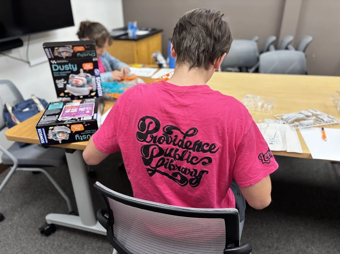 A person with short brown hair wearing a pink t-shirt with black text that reads "Providence Public Library" is sitting at a table assembling a toy kit. In the background, a young girl is also working on a craft project.
