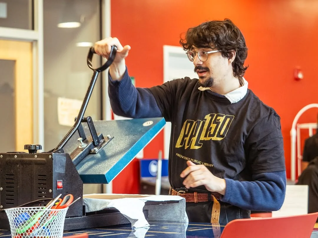 A young man with curly brown hair, glasses, and a beard peeling a vinyl record at a library or community center.
