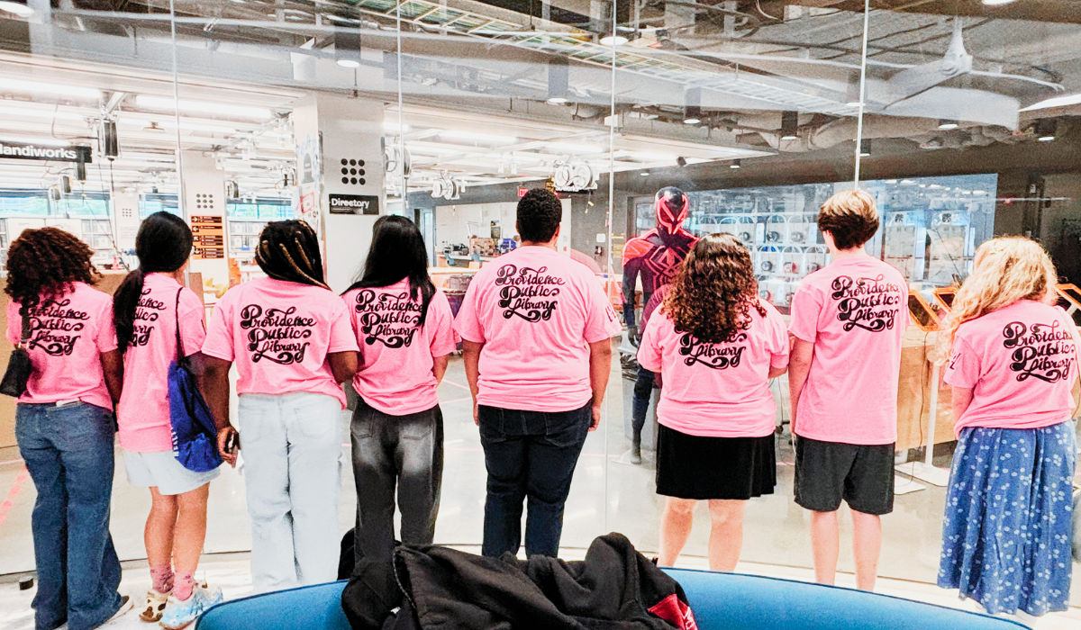 Group teens form PPL STEAM summer, wearing matching pink t-shirts with 'Providence Public Library' printed on the back while looking through Northeastern University Makerspace