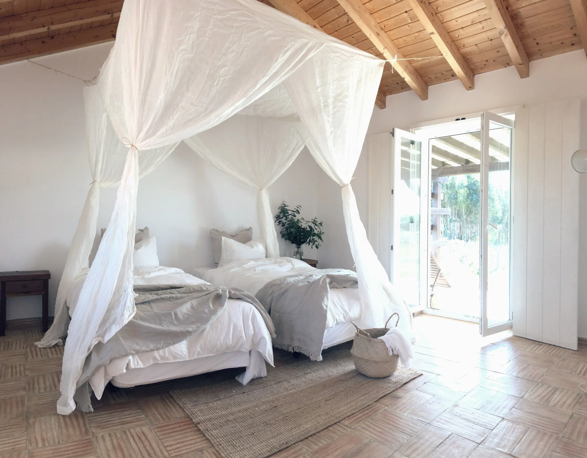 A cozy bedroom with white bedding and a canopy bed in a room with wooden beams and a door opening to a porch with outdoor views.