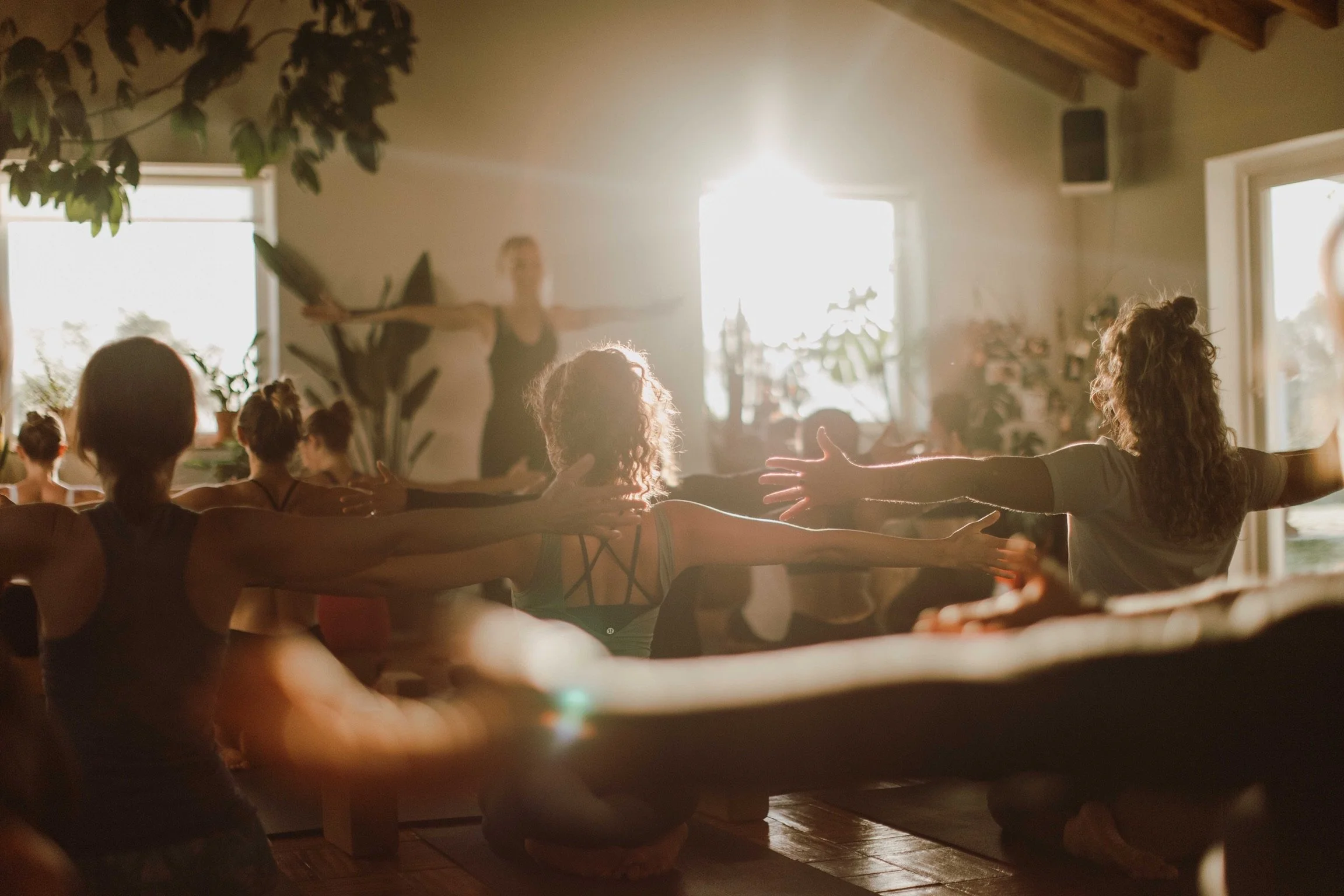 People participating in a yoga or meditation class in a sunlit room, sitting cross-legged with arms extended outward, following an instructor at the front.