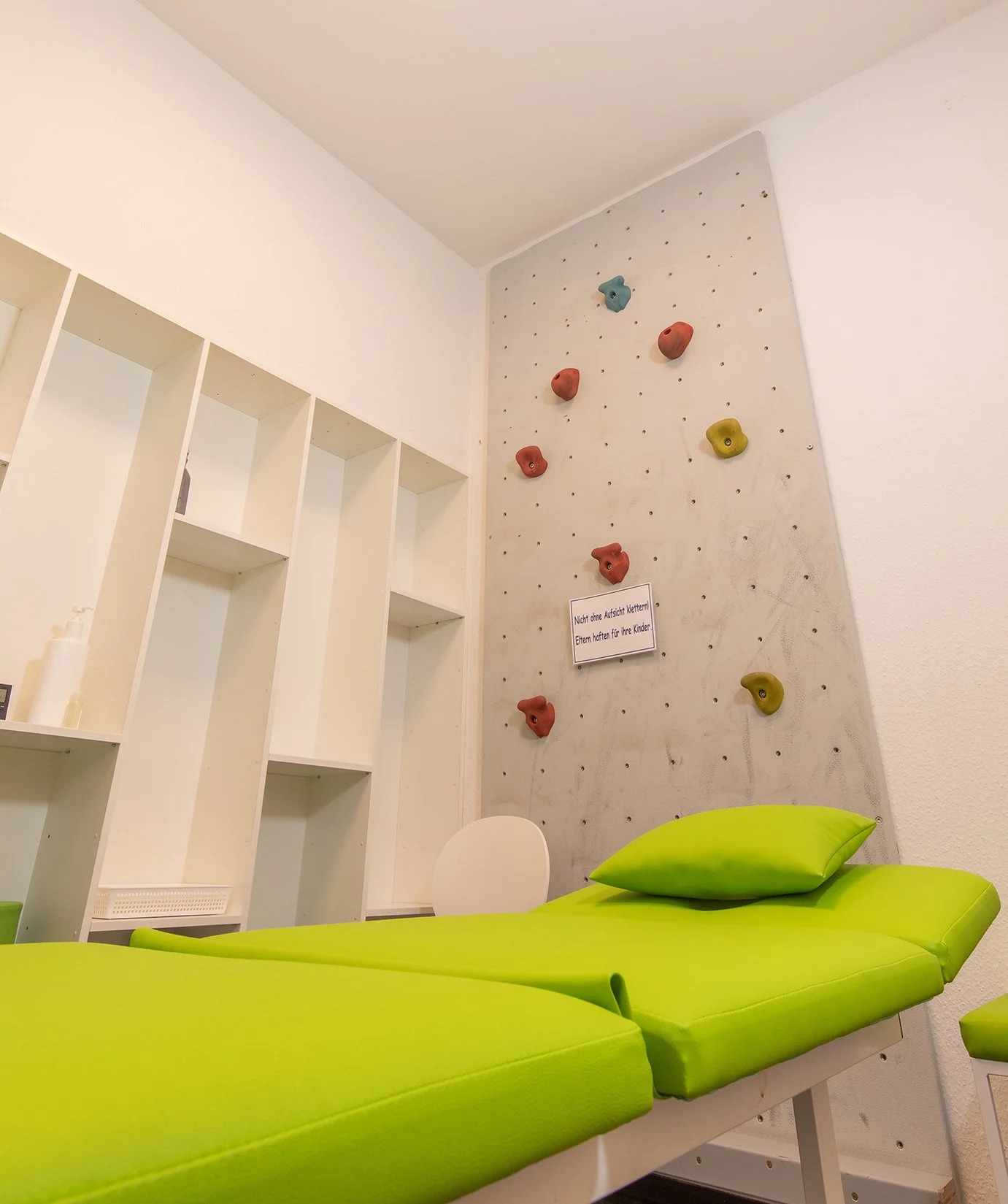 Eine Krankenzimmer mit einer grünen Liege, weißen Wänden, einem Kletterwand mit bunten Griffen und einem Schild, das vor Aufsicht warnt.
