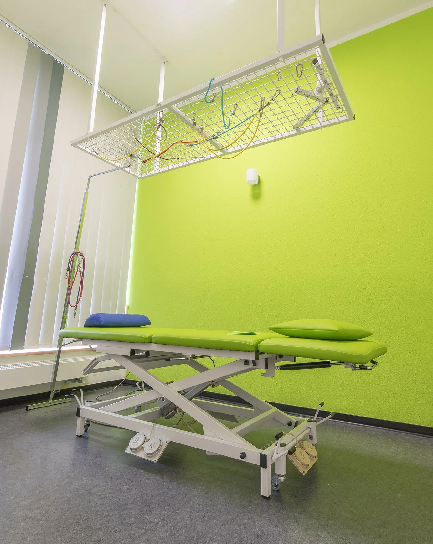 Behandlungsraum mit Untersuchungsliege in grün und vertikaler Lamellenfenster, Wand in hellgrün.