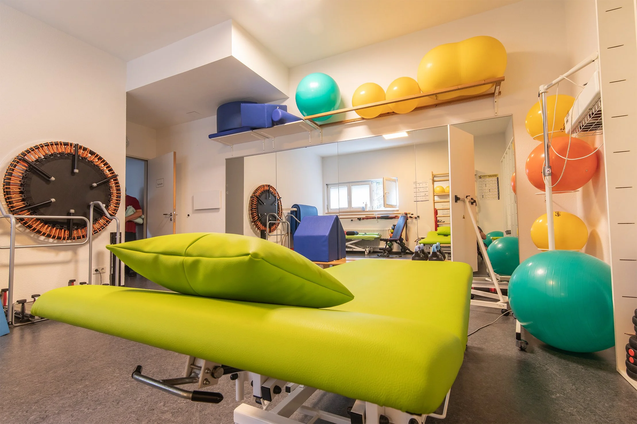 Fitnessraum mit grüner Liege, buntenGymnastikbällen, Spiegel, Trainingsgeräten und Fenstern.
