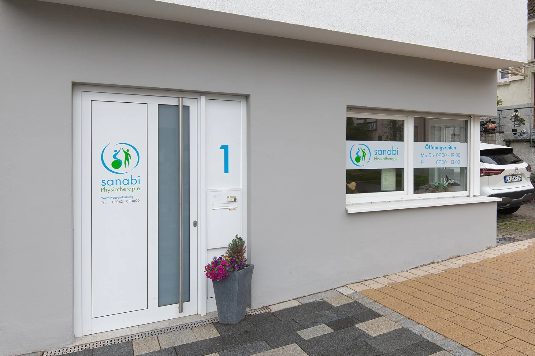 Außenansicht einer Physiotherapie Praxis namens 'sanabi' mit weißen Türen und Fenstern, geöffneten Öffnungszeiten, einer Blumenpflanzung vor der Tür und einem Parkplatz im Hintergrund.