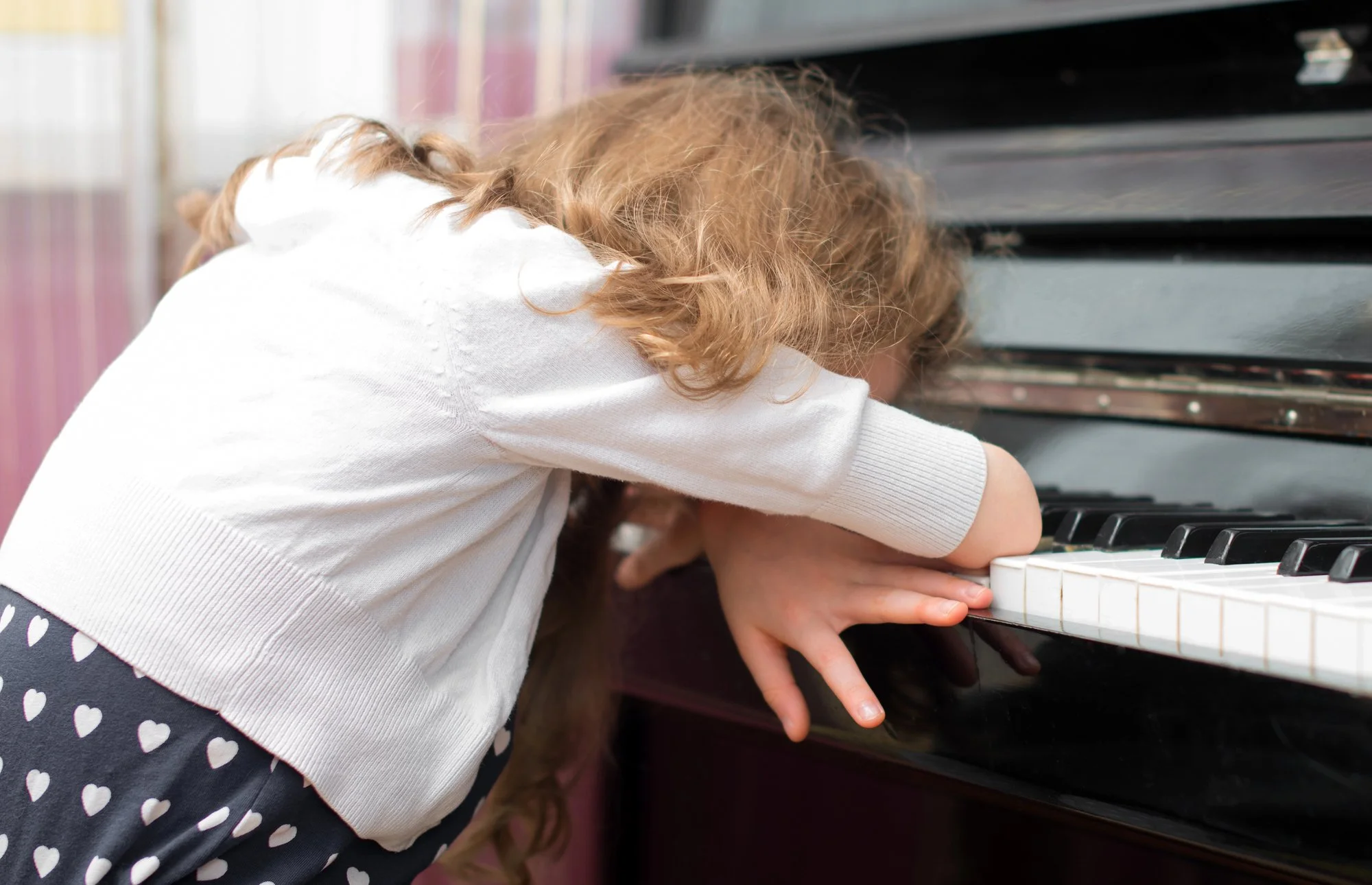 A young girl with curly hair leaning over a piano keyboard, possibly playing or looking at the keys.