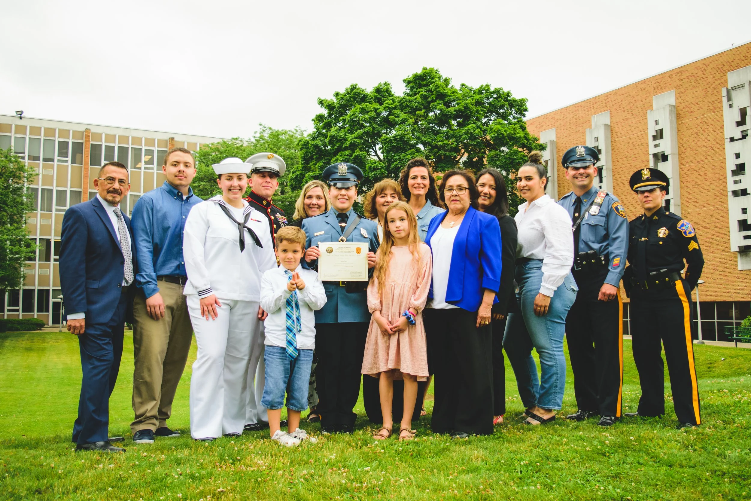 Group of police officers, military personnel, children, and adults standing on grass in front of a school building, celebrating a certificate award.