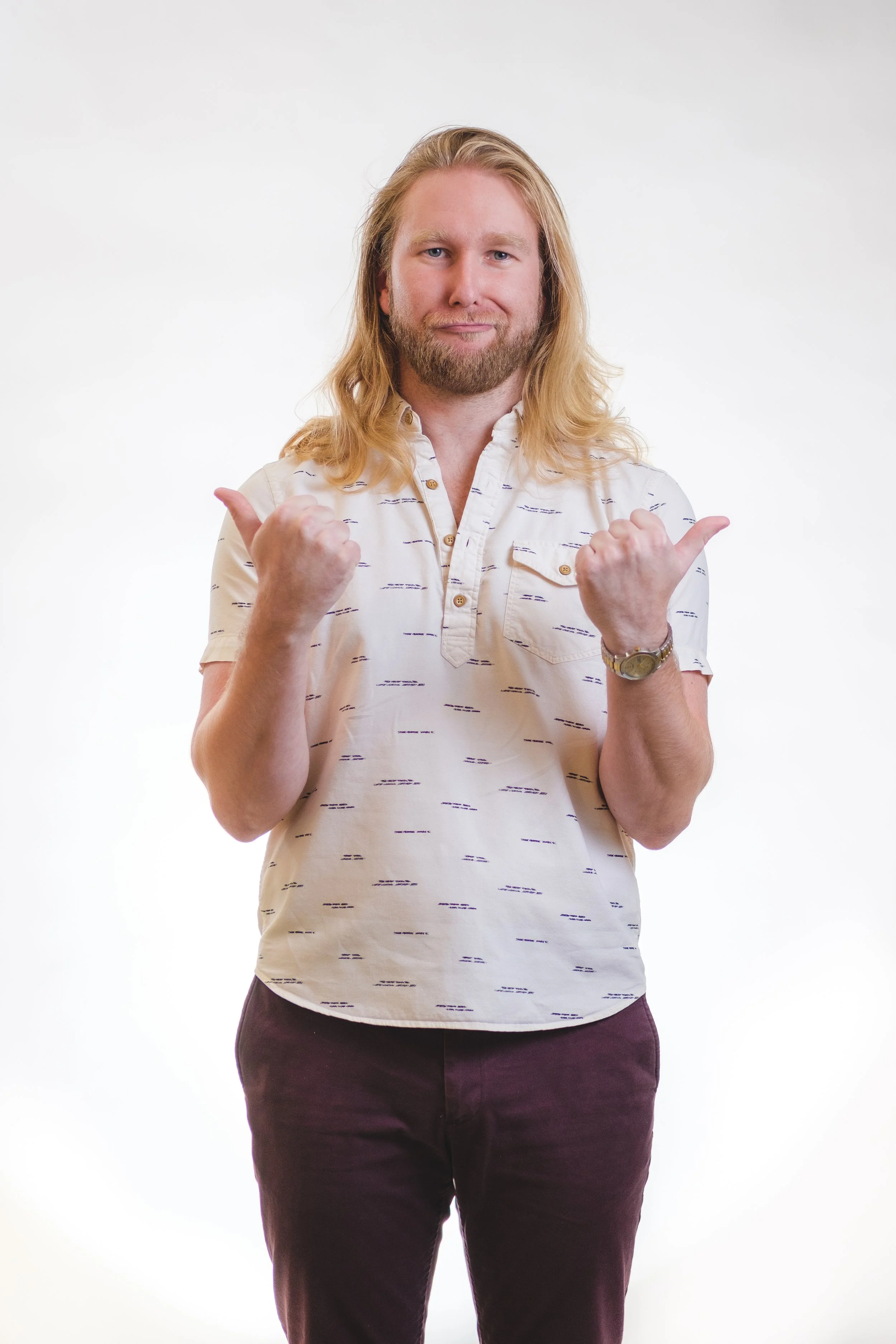 A man with long blond hair and a beard, wearing a light-colored patterned shirt and dark pants, is standing and showing a thumbs-up with both hands.