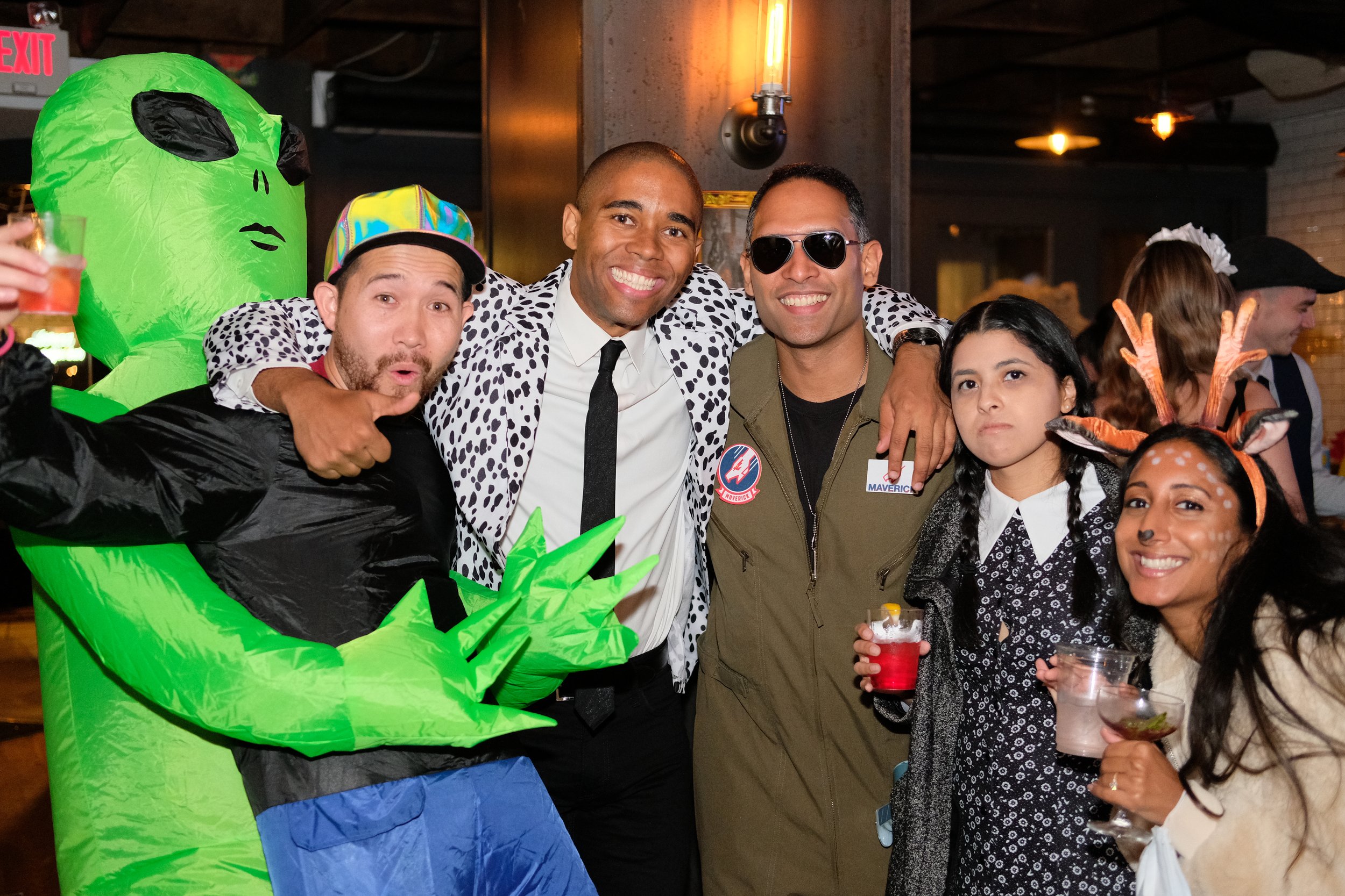 Group of five friends at a party, dressed in festive and costume attire, holding drinks and smiling, with a large inflatable green alien in the background.