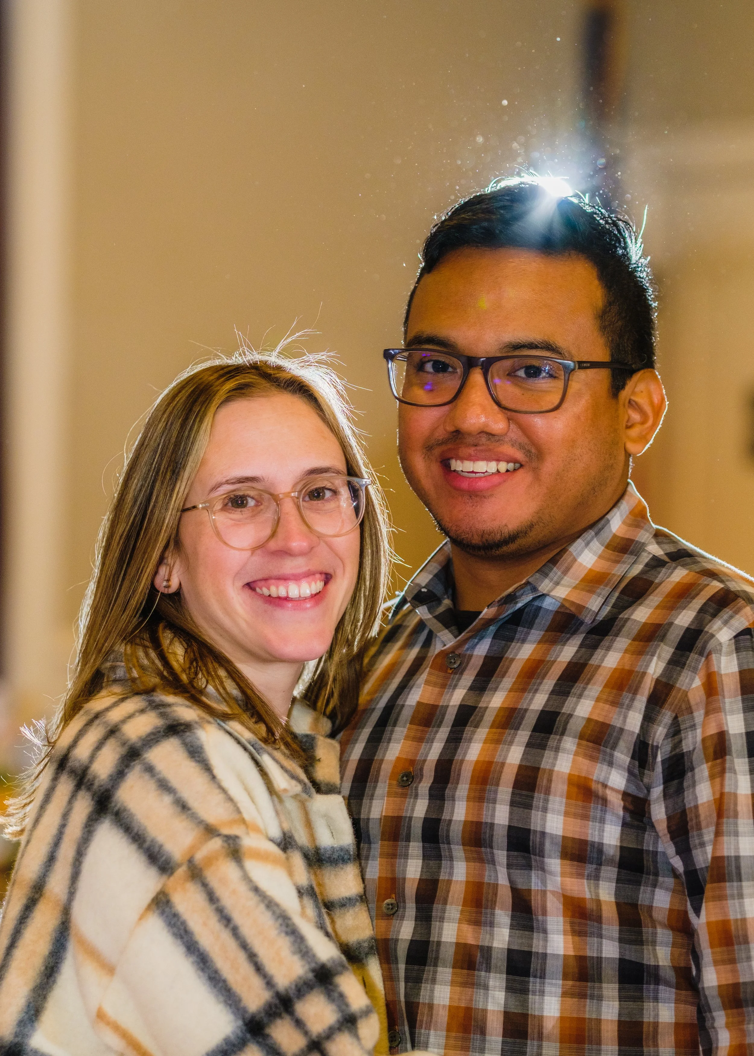 A smiling couple with glasses standing close together in an indoor setting, with a light source behind them creating a glow around their heads.