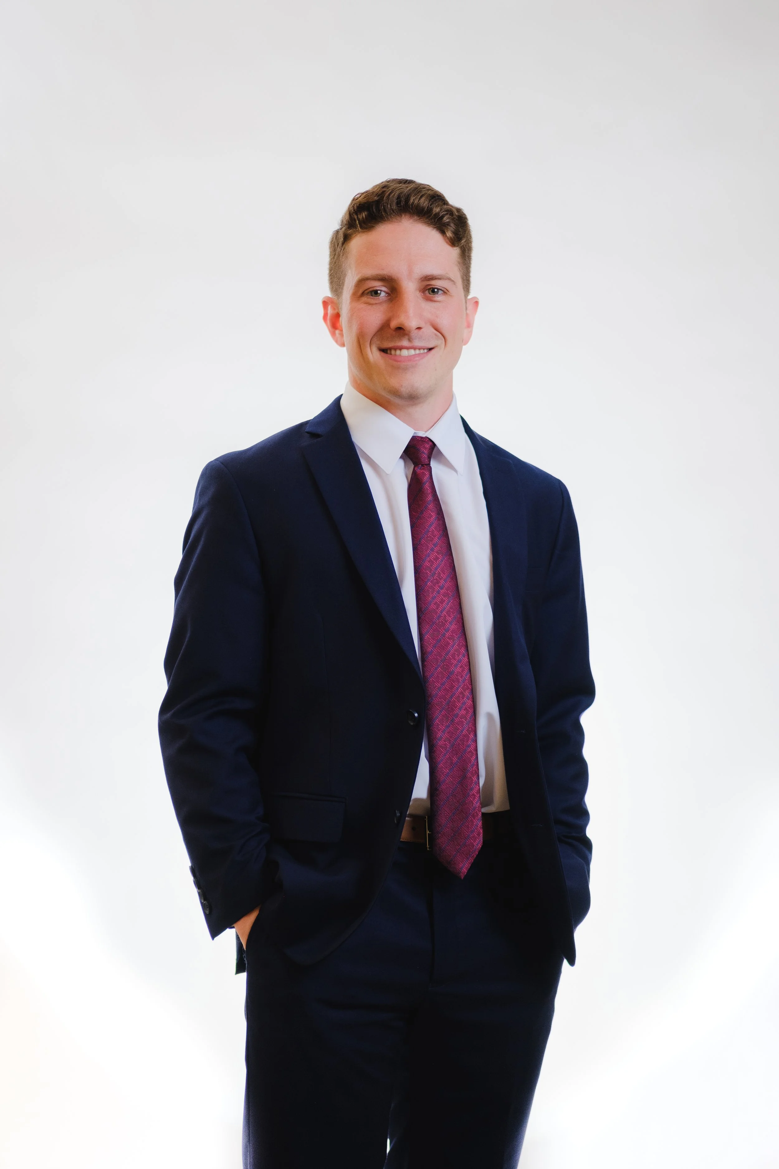 A young man with short light brown hair, wearing a navy blue suit and a white shirt, with a red patterned tie, standing against a plain white background and smiling at the camera.