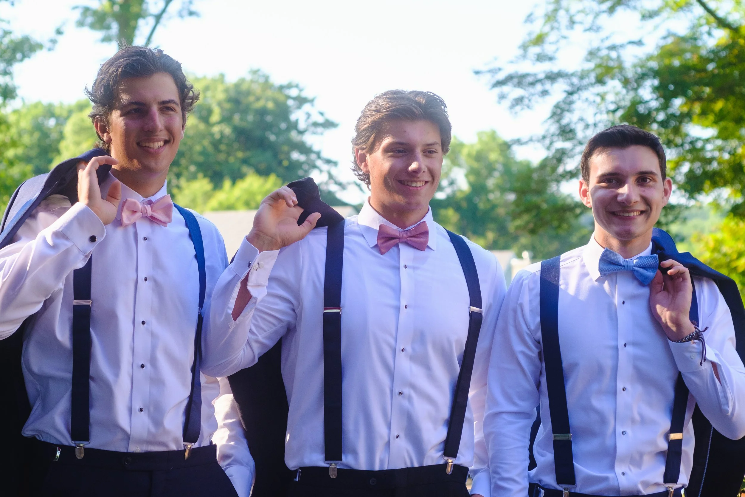 Three young men in white shirts with suspenders and bow ties smiling outdoors, holding jackets over their shoulders, with green trees in the background.