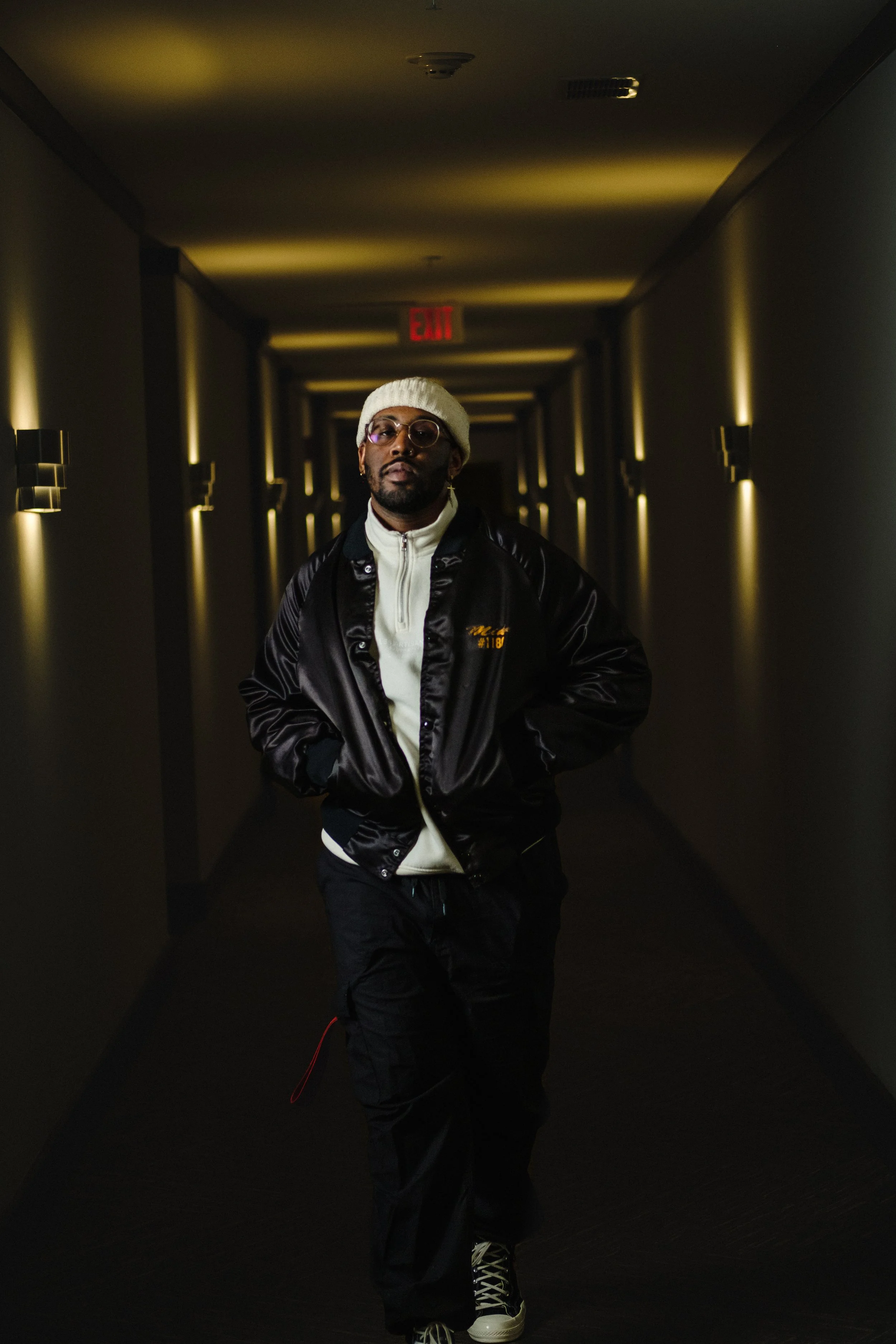A man walking down a dimly lit hallway with modern light fixtures on the walls, wearing a white knit beanie, glasses, a black satin jacket, a white shirt, black pants, and sneakers.