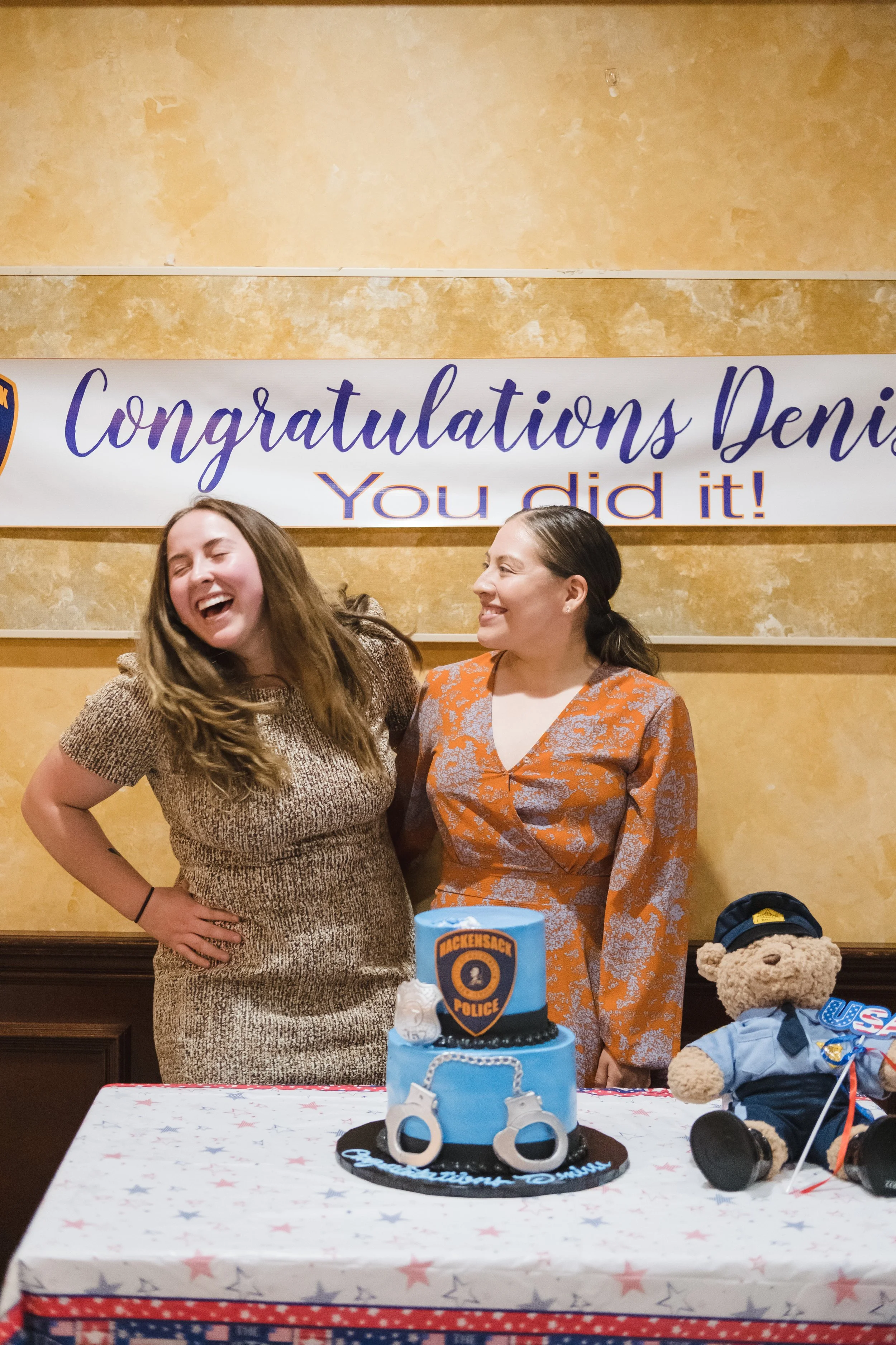 Two women happy and smiling at a police-themed graduation celebration, with a cake decorated with police badges and handcuffs, and a banner in the background reading, 'Congratulations Deni, You did it!'