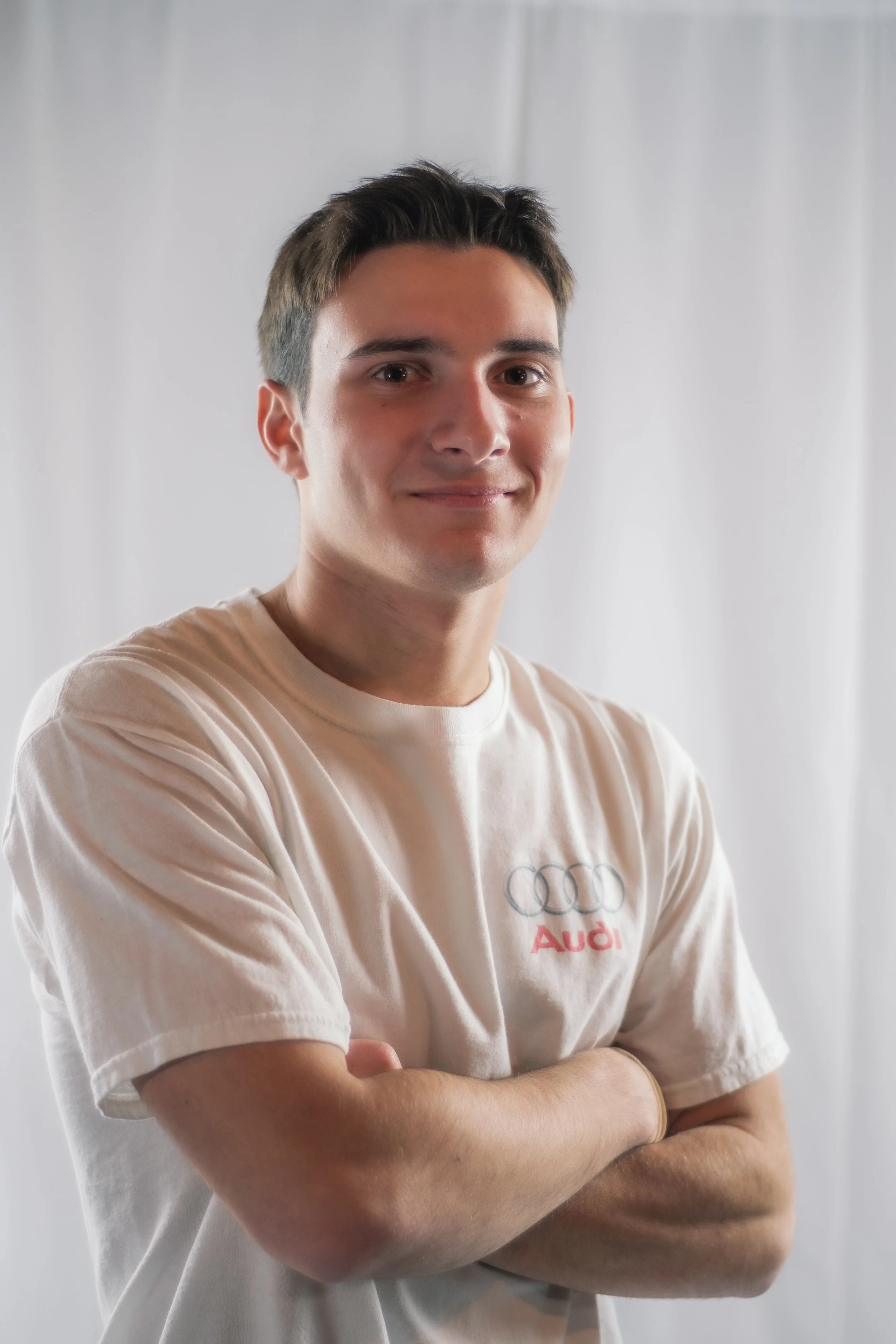 A young man with short brown hair, wearing a white T-shirt with Audi logo, standing with arms crossed, smiling against a white background.