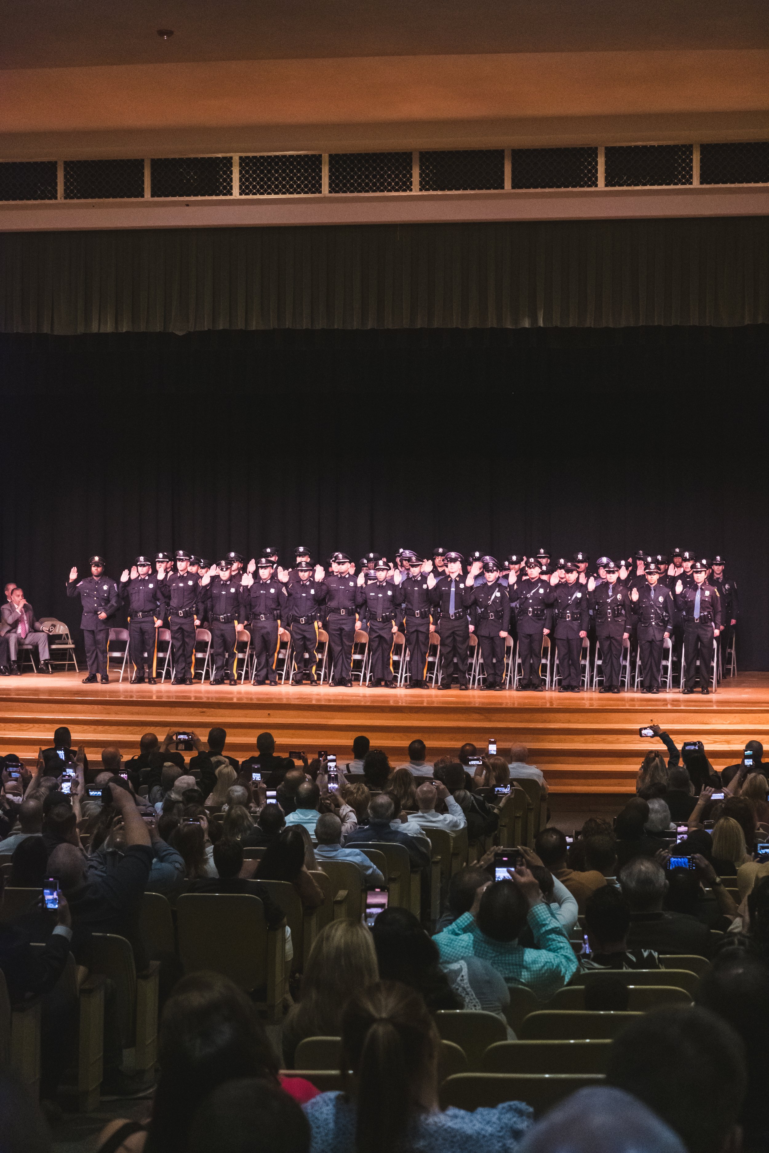 Group of police officers on stage during a ceremony or event, with audience members taking photos.