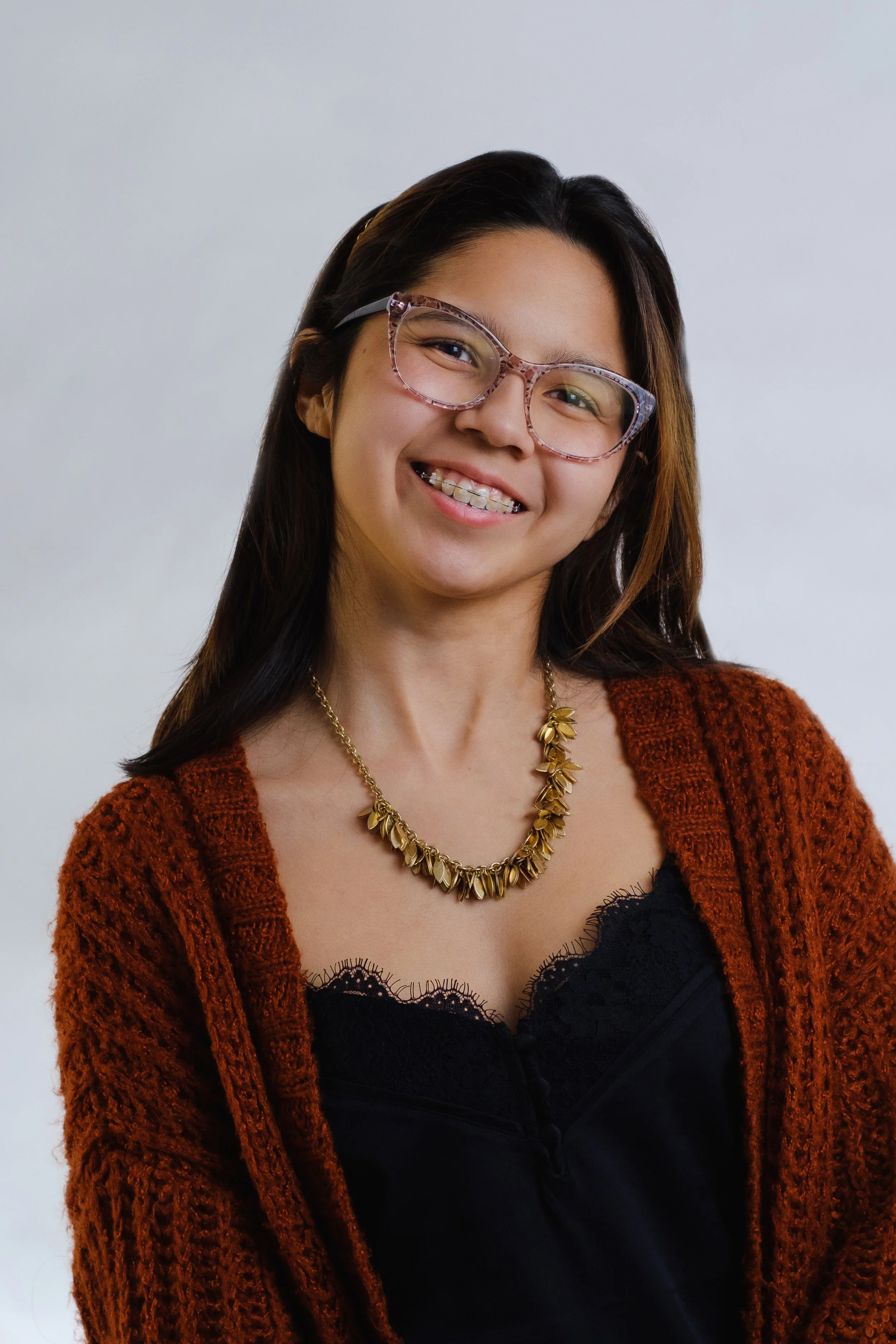 A smiling woman with glasses, braces, wearing a black top, a rust-colored cardigan, a gold necklace, and a headband, standing against a plain background.