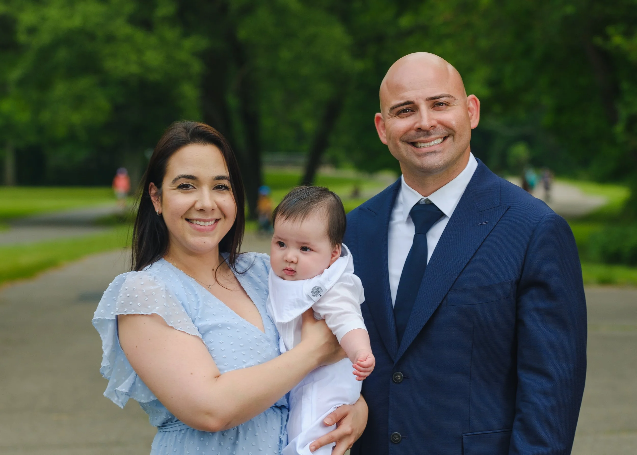 A smiling woman with dark hair holding a baby dressed in white, standing next to a smiling man in a blue suit outdoors in a park with green trees and a path in the background.