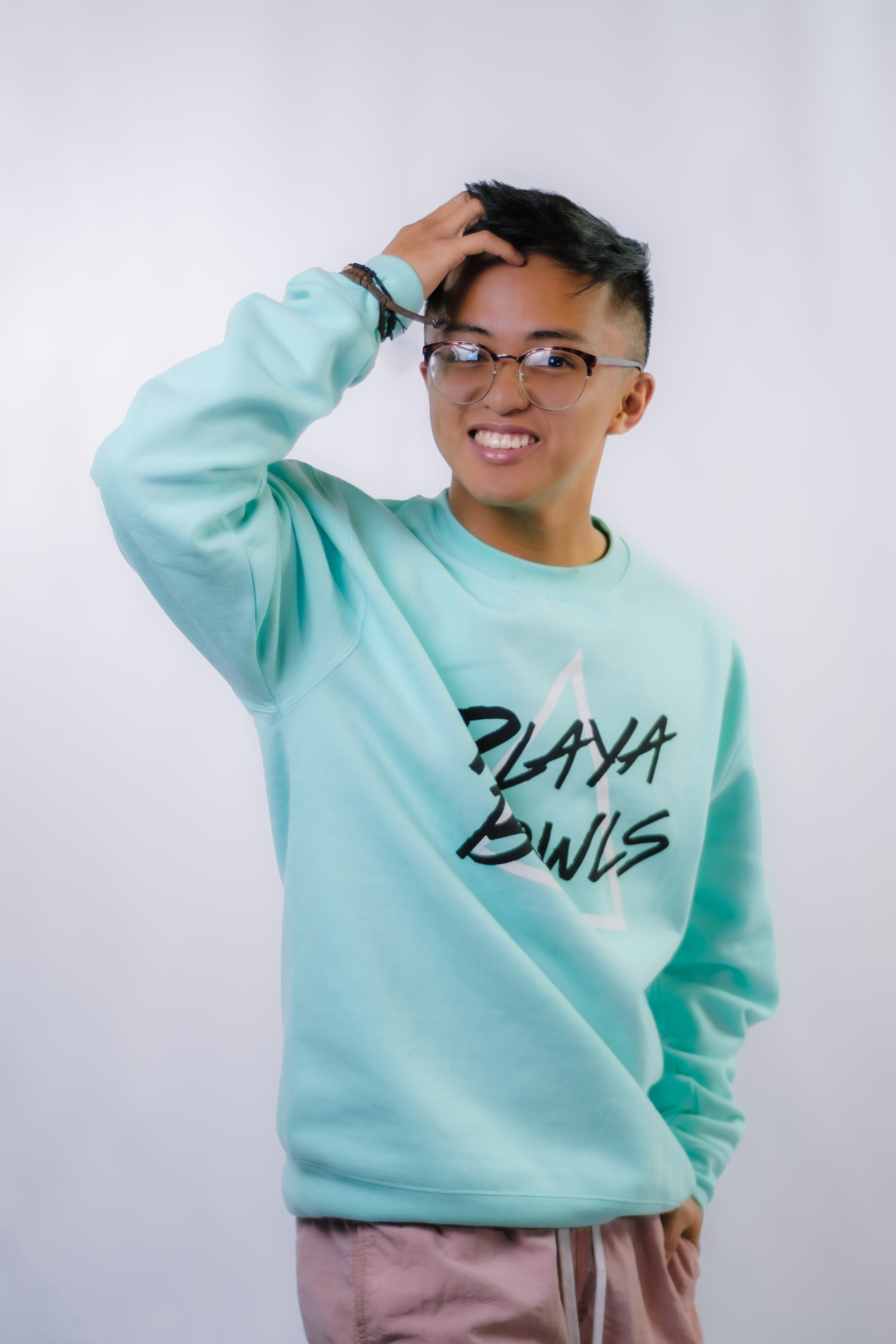 A young man with glasses, wearing a turquoise sweatshirt with black and white text, has black hair and is touching his head with one hand, smiling against a plain background.