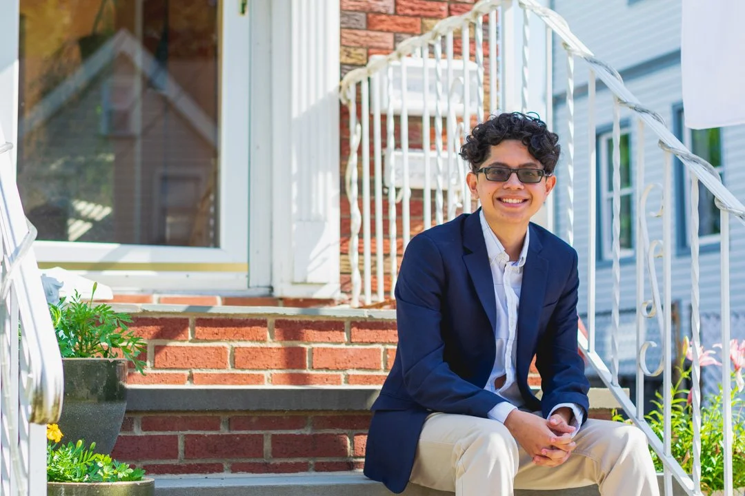 Young man in a blue blazer and beige pants sitting on front porch steps, smiling, with plants and house details in the background.