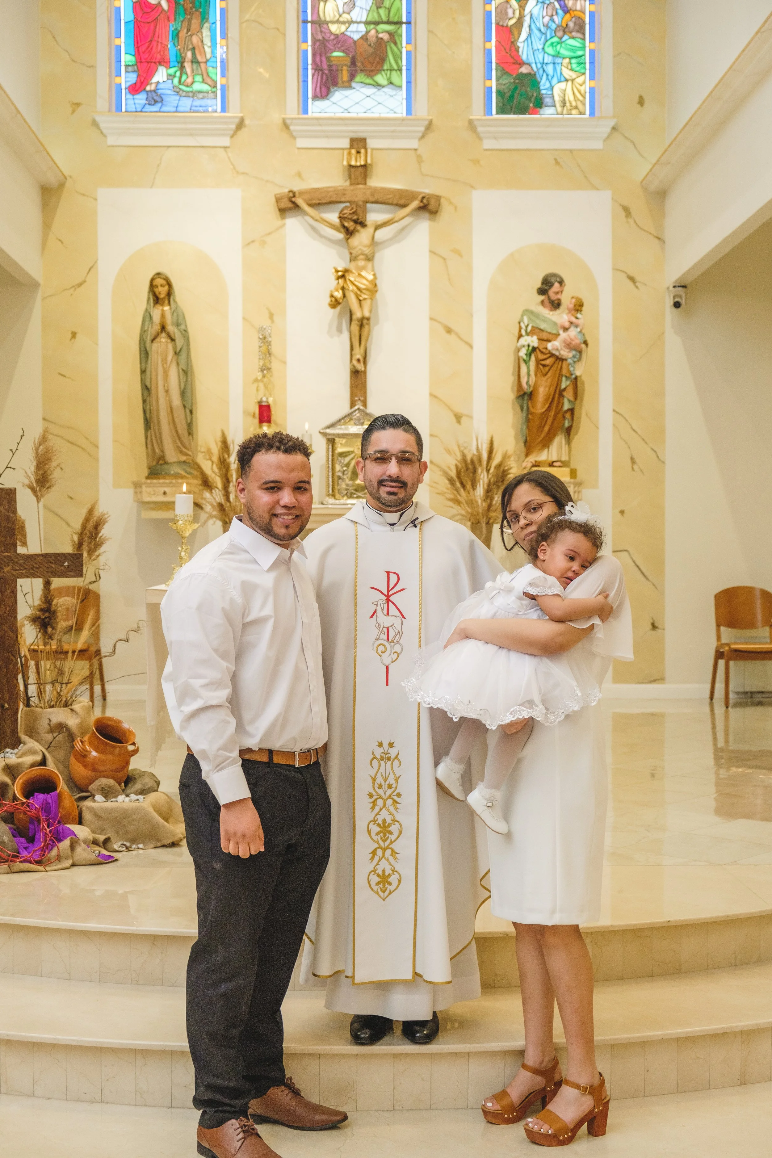 A family is posing with a priest inside a church during a religious ceremony, likely a baptism, with a decorated altar and religious statues and images in the background.