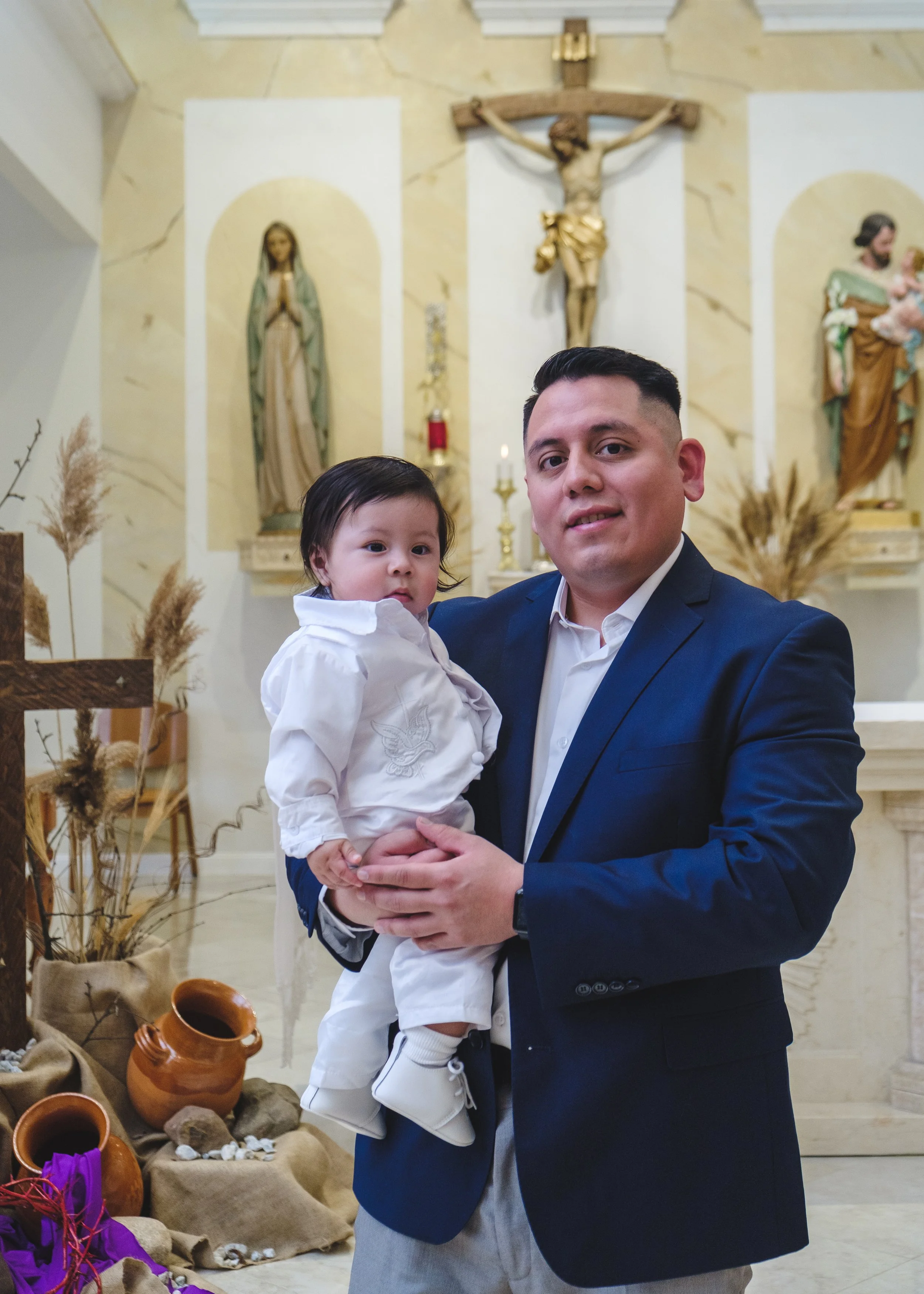 A father in a navy blue suit holding a young child dressed in white inside a church with religious statues and decorations in the background.