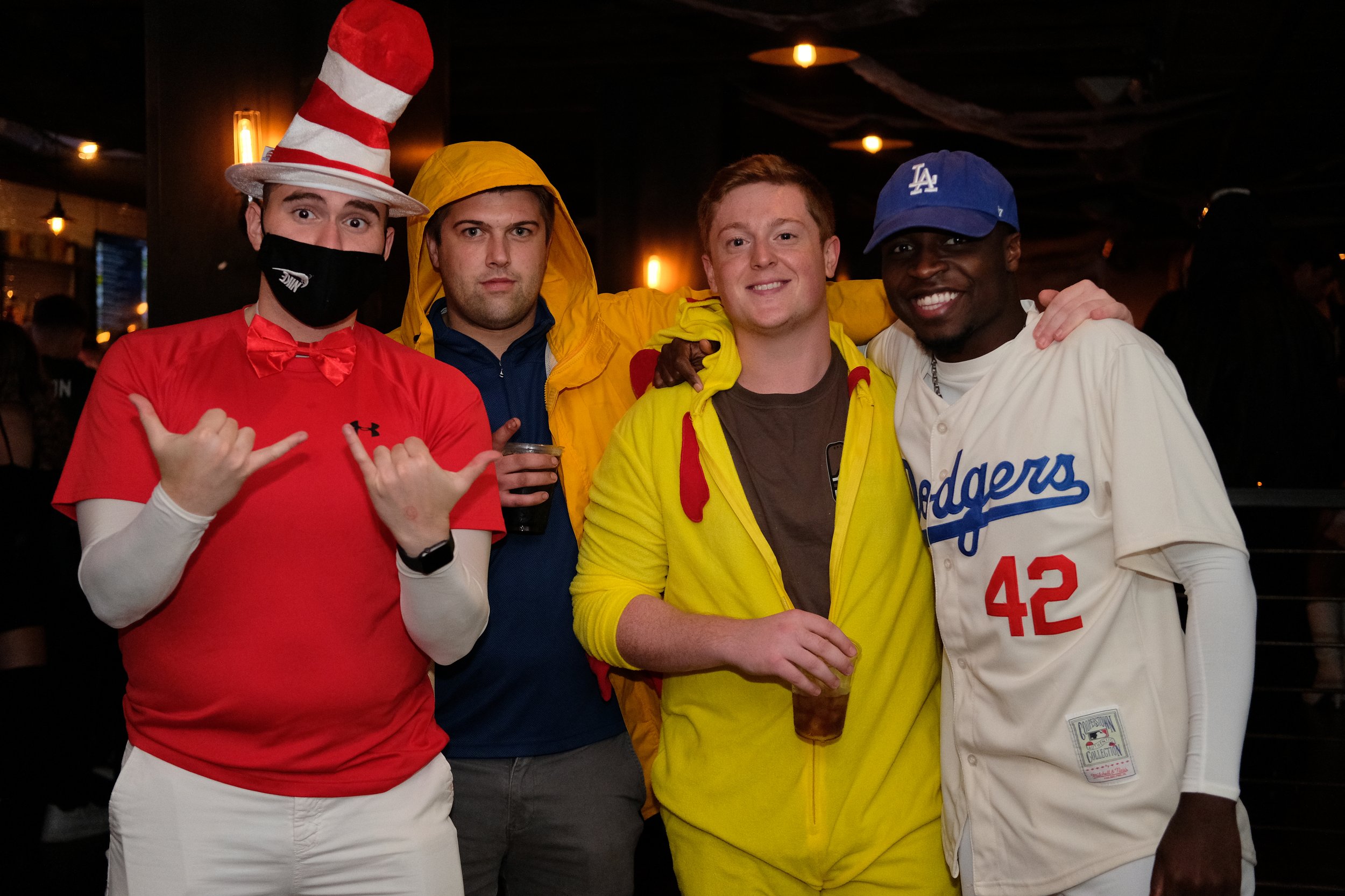 Four men dressed in colorful casual and sports outfits, posing together at a social event or party with warm lighting.
