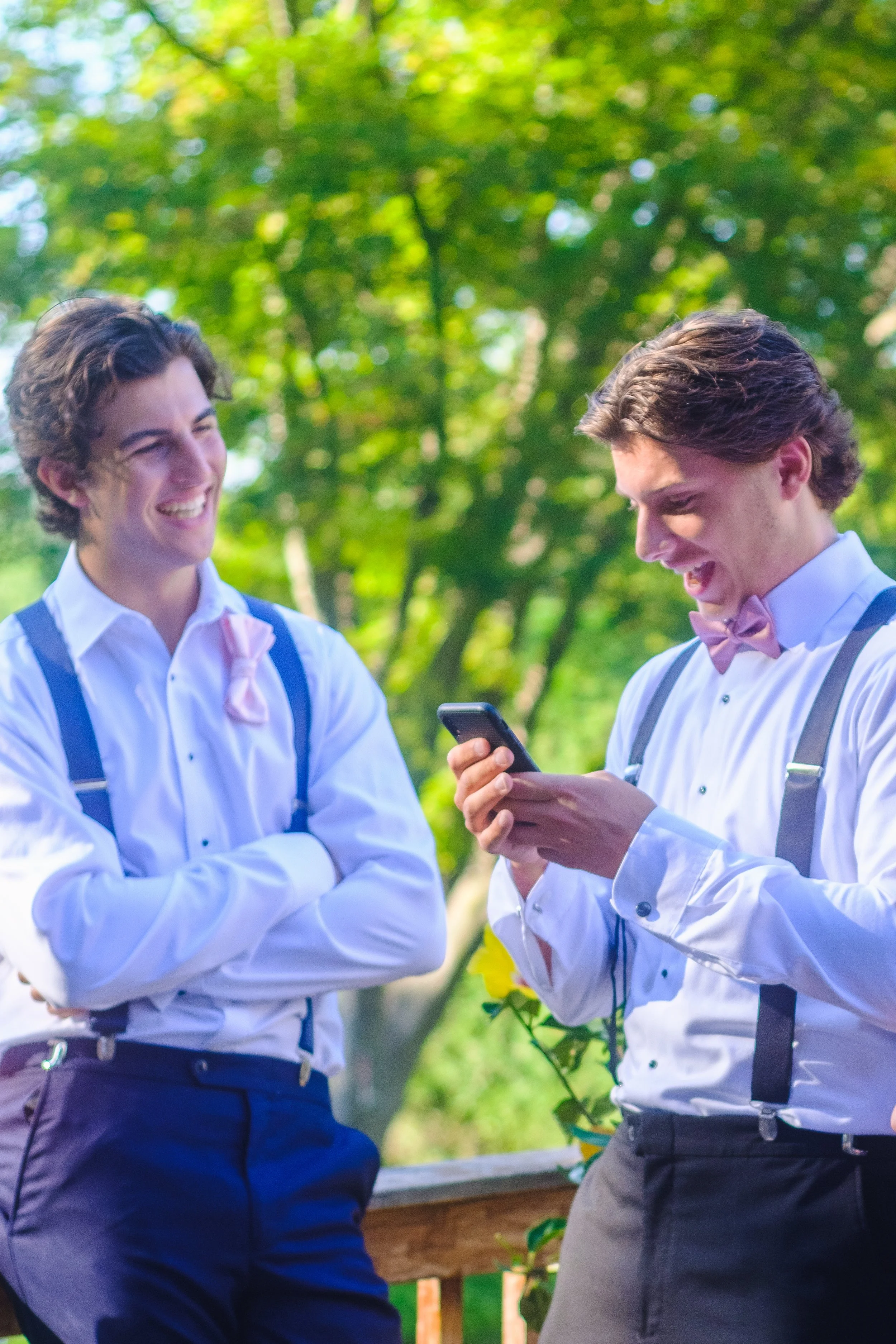 Two young men dressed in formal attire with suspenders and bow ties laughing and looking at a smartphone outdoors in a green park.