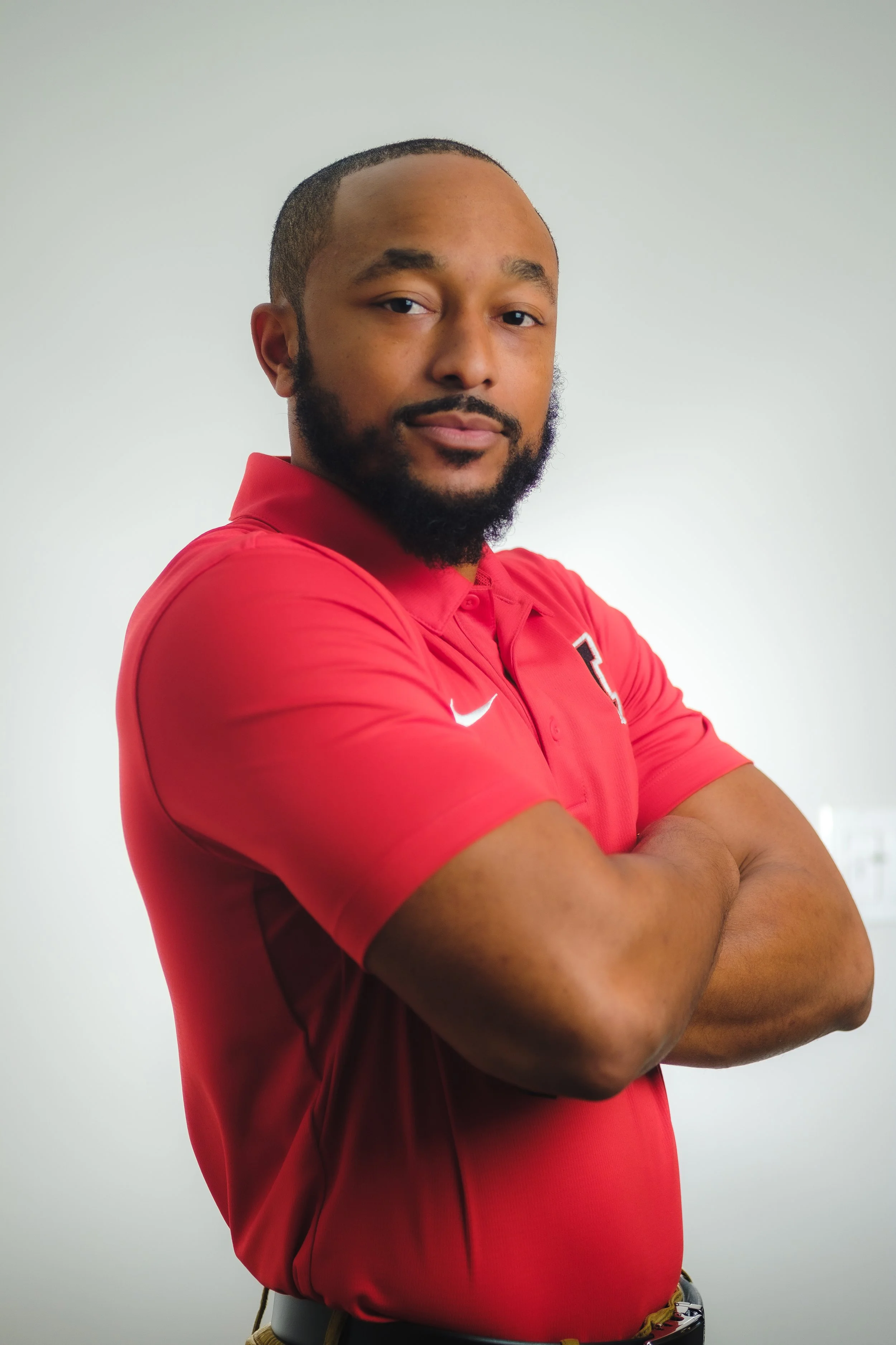 Portrait of a man with a beard, wearing a red collared shirt with a Nike logo, crossing his arms against a plain white background.