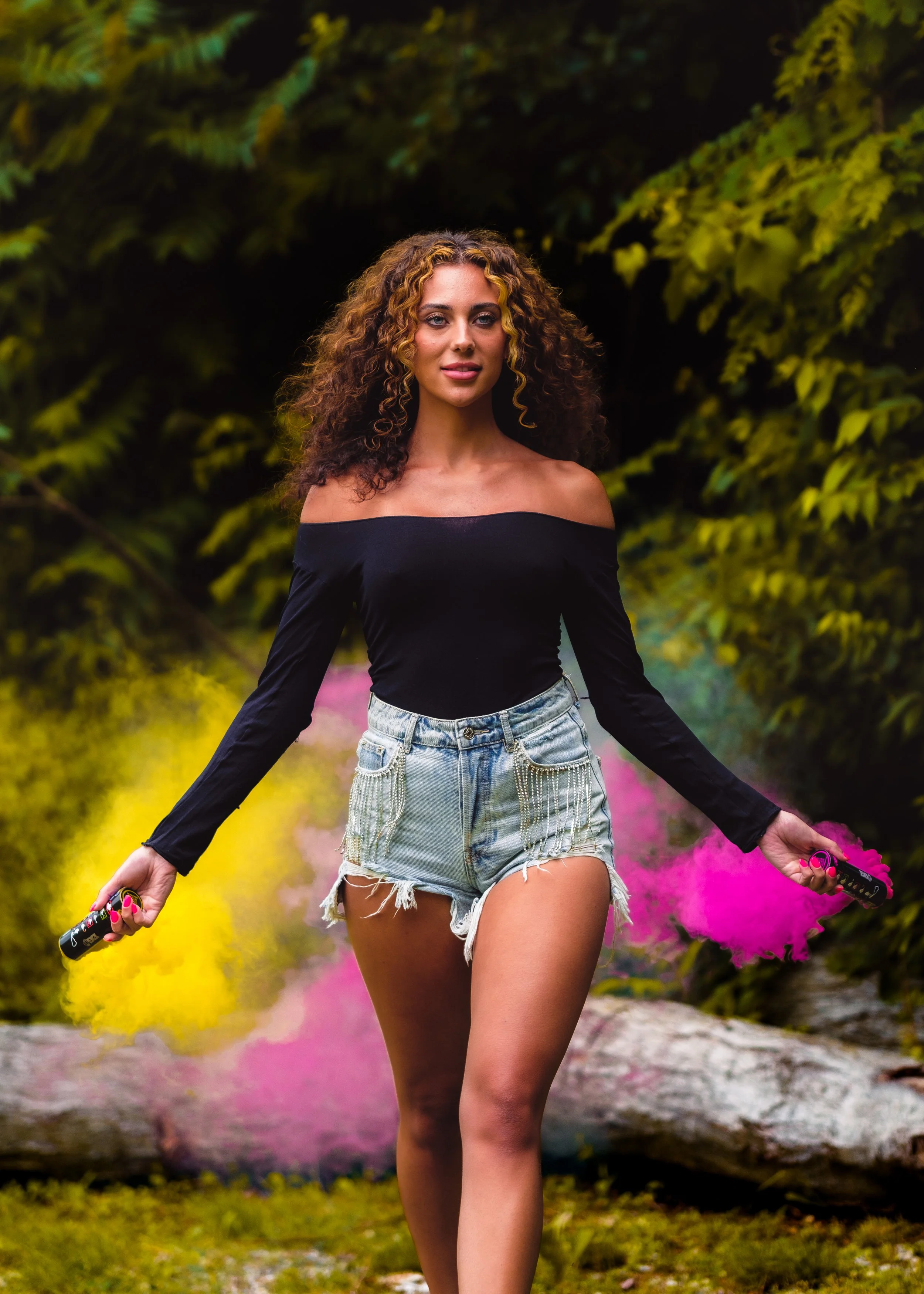 Young woman with curly hair walking outdoors holding colorful smoke emitters, surrounded by green foliage.